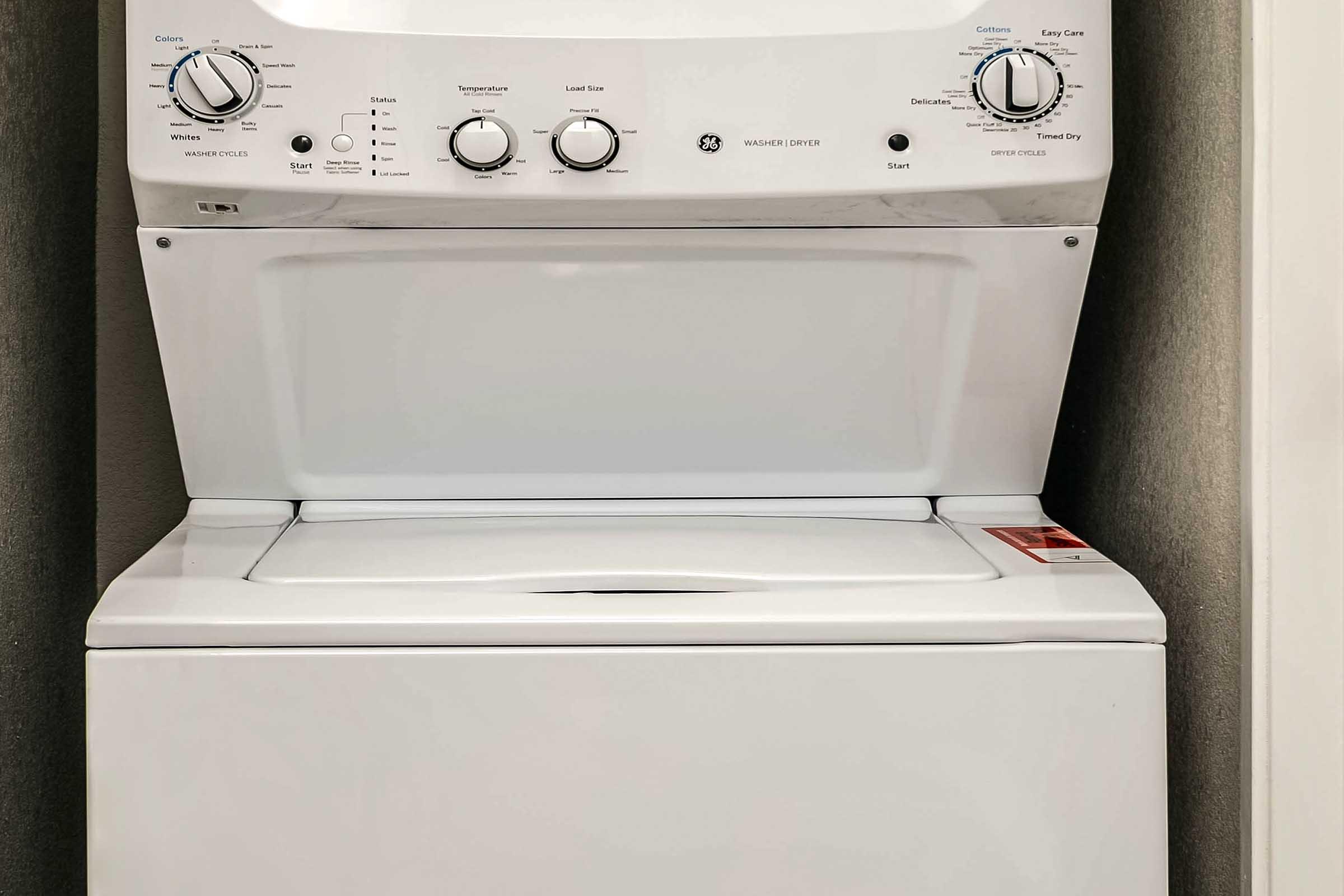 A white washer and dryer combo unit, featuring control knobs and a digital timer on the front. The machine is positioned against a wall, and the top section is flat, suitable for placing items. The design is modern and compact, ideal for small laundry spaces.