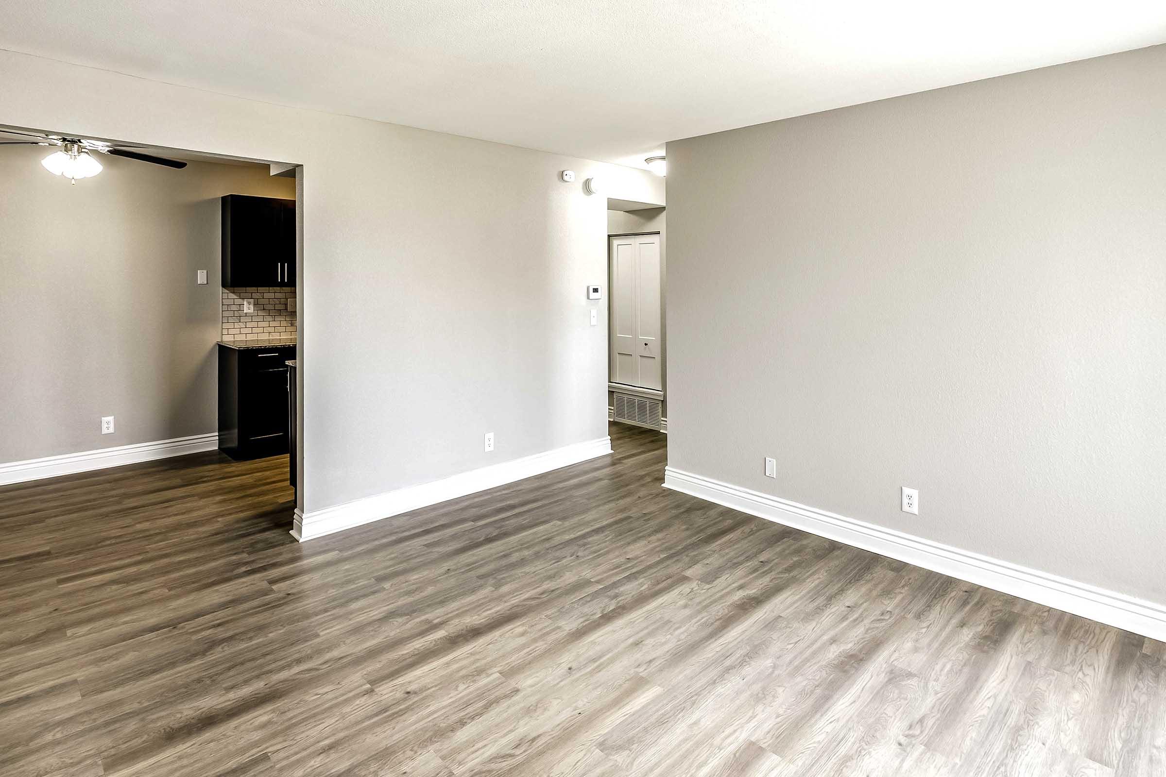 A bright, unfurnished room with light gray walls and a wood laminate floor. To the left, there is a doorway leading to a kitchen area with dark cabinetry. The space is well-lit, featuring a ceiling fan, and has an open layout leading to another area.
