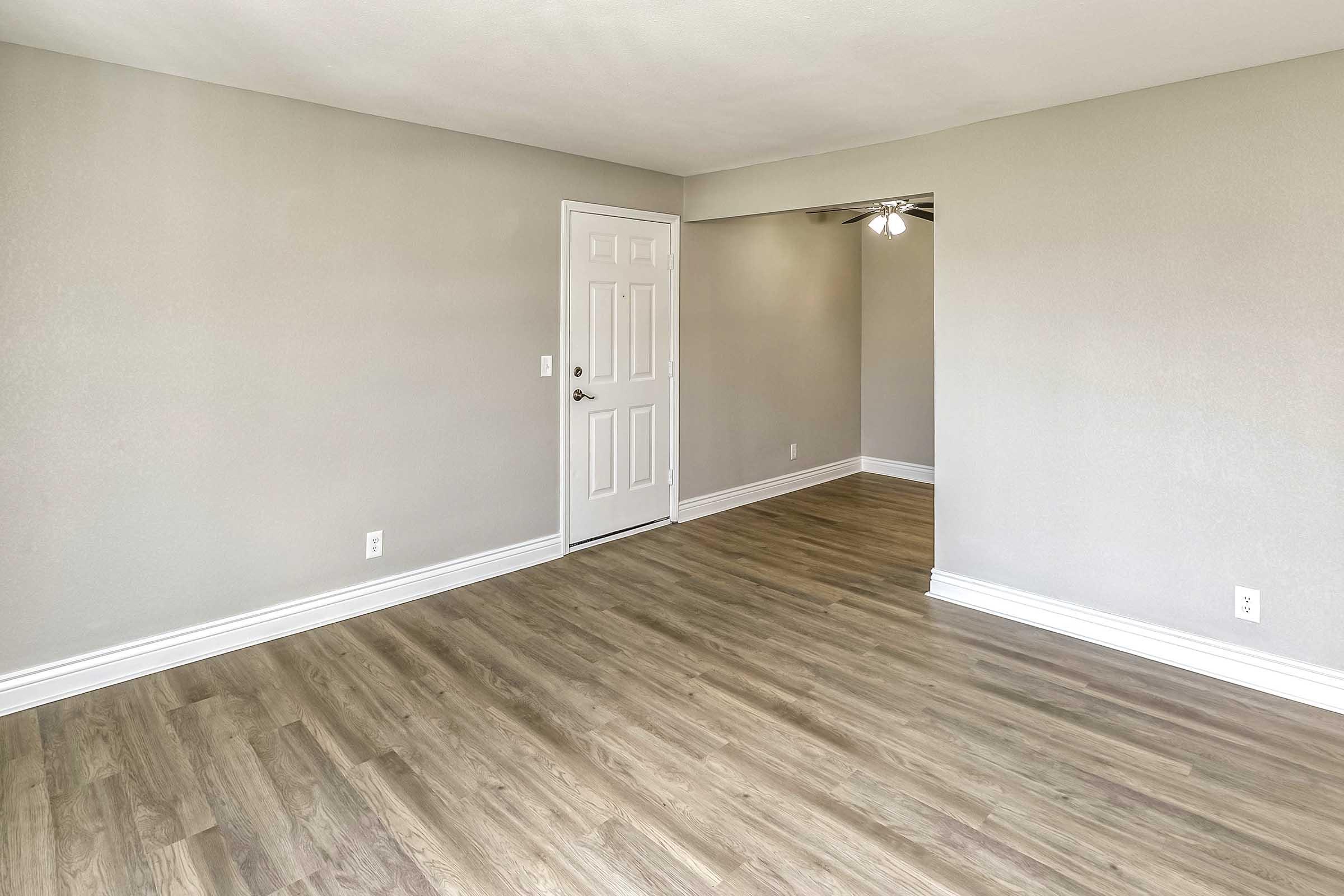 A spacious, empty room with light gray walls and a light wood floor. There is a white door on the left side, leading to another room. A ceiling fan is visible in the background, and the overall ambiance is bright and modern.