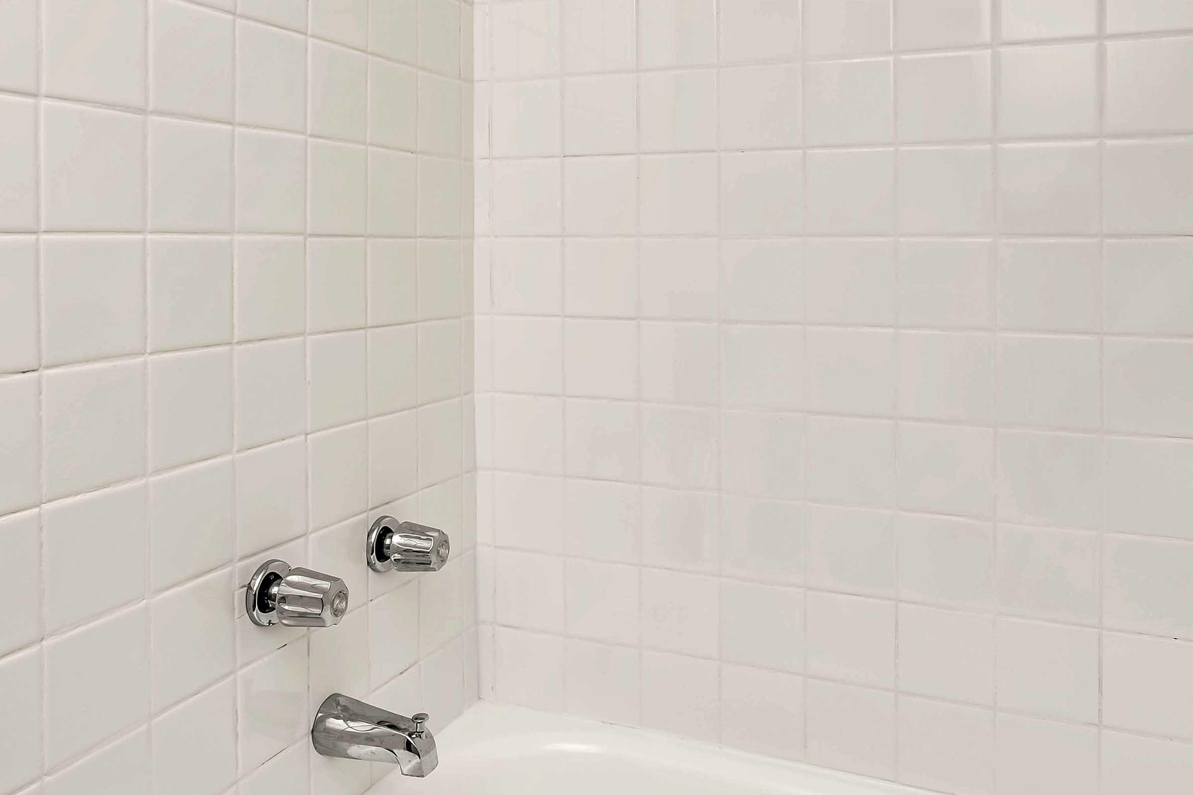 A clean, white tiled bathroom wall with silver faucets and shower controls. The tiles are arranged in a grid pattern, creating a bright and modern appearance. The bathtub is partially visible, adding to the bathroom's simplicity and functionality.