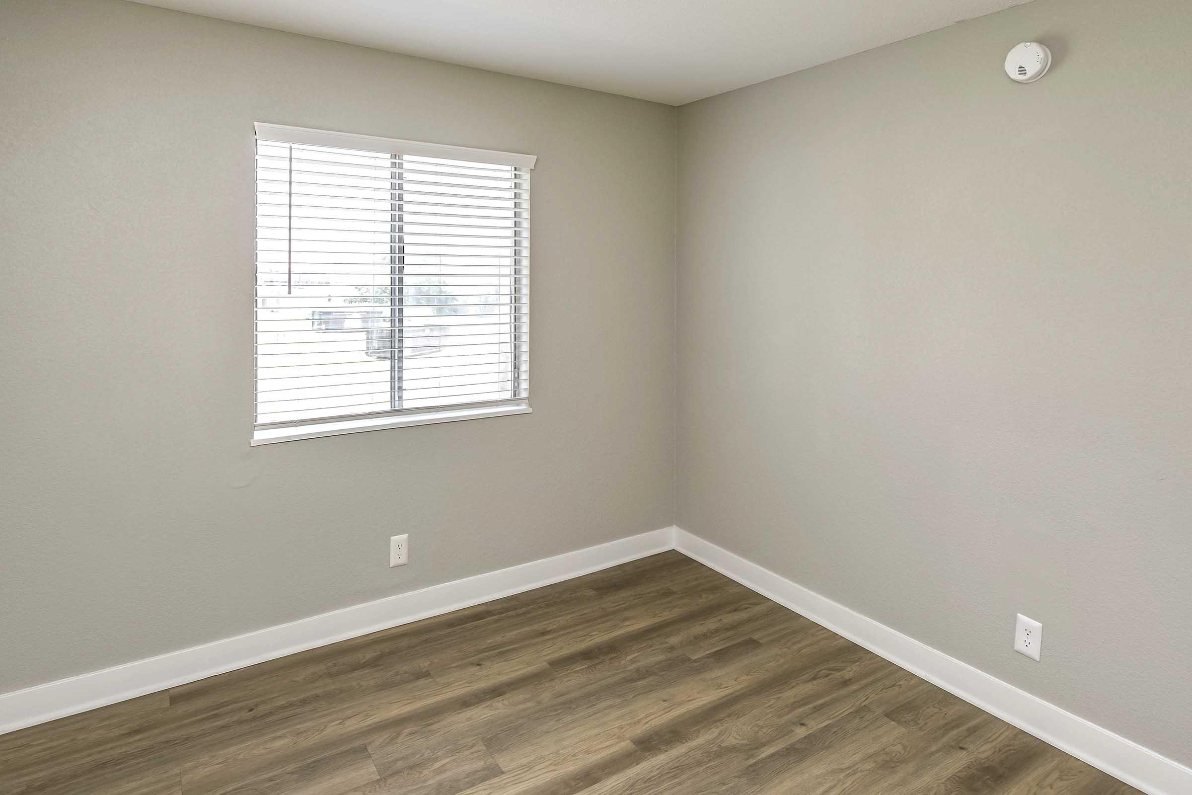 A minimalistic room featuring light gray walls and a window with blinds. The floor is covered in light-colored laminate, and there is a small white smoke detector on the ceiling. The space is empty, creating a clean and spacious atmosphere.