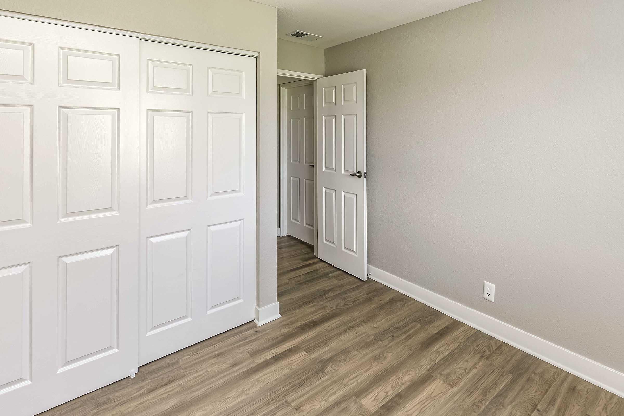 Interior view of a room featuring light gray walls and wooden flooring. There are two sets of closed white double doors on the left and an open door leading to another area on the right. The space is well-lit and minimalist, creating a modern and spacious feel.