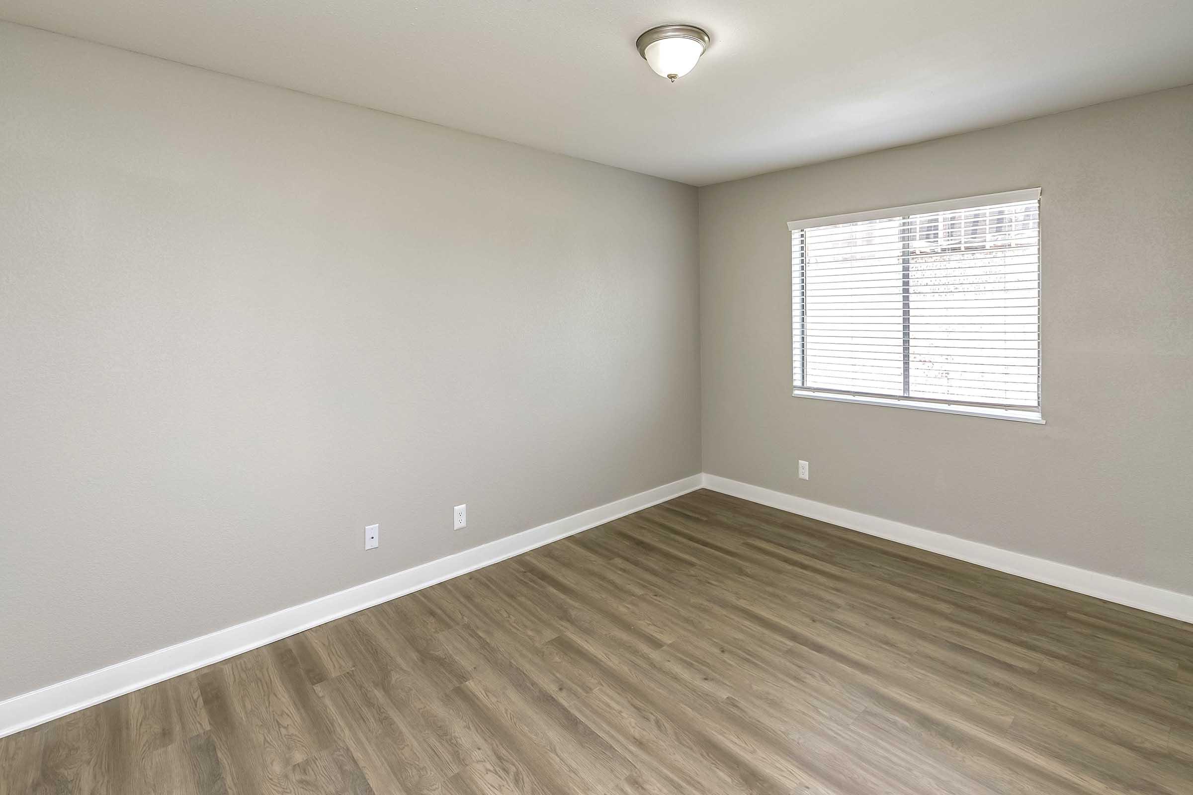A well-lit, empty room with smooth, light-colored walls and a wood-like floor. There is a ceiling light fixture and a window with blinds. The room has no furniture, creating an open and spacious feeling.