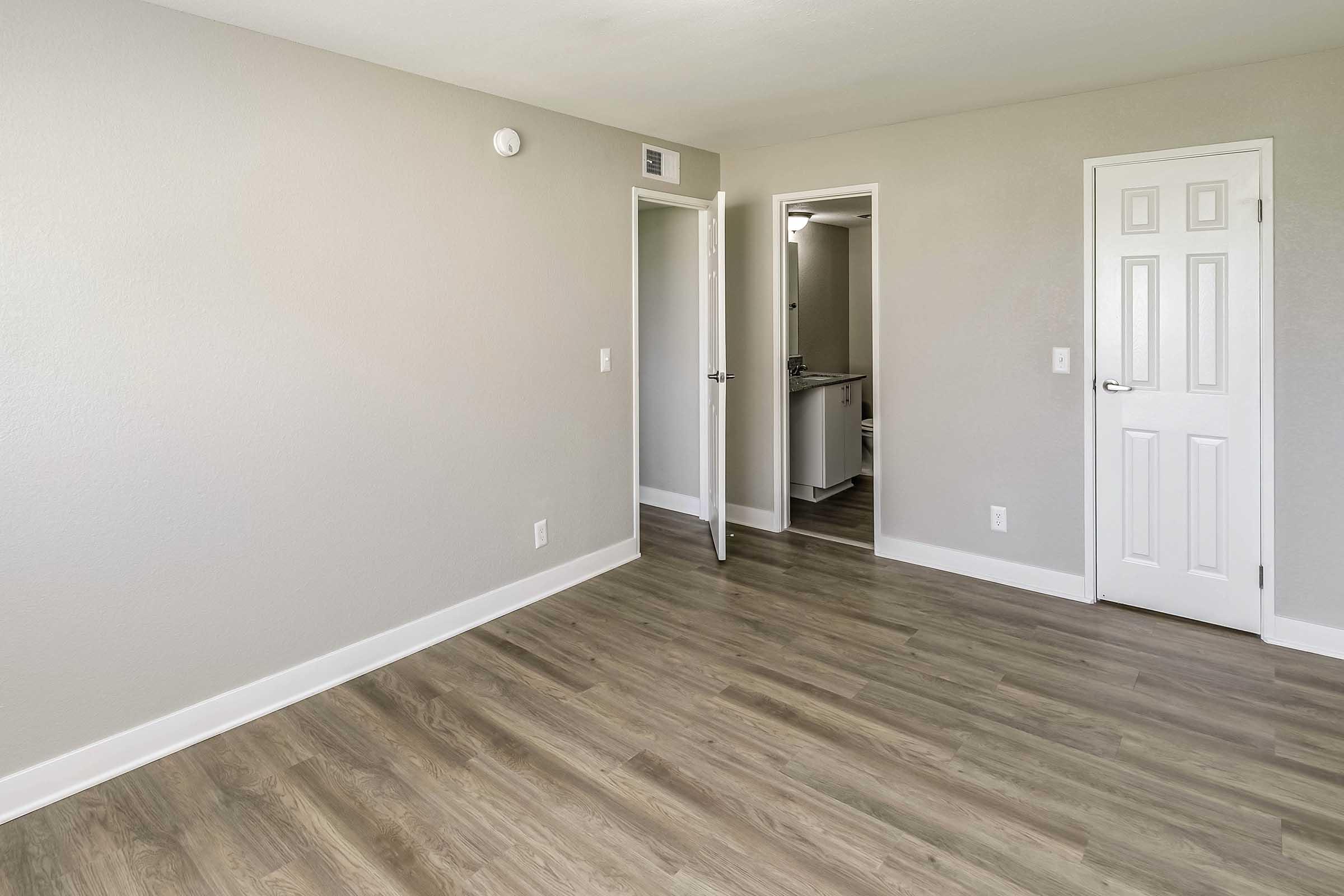 A spacious, empty room with light gray walls and hardwood-style flooring. A closed white door is visible on the right, and another door leads to a small bathroom area in the background. Natural light fills the space, creating a bright and airy atmosphere.