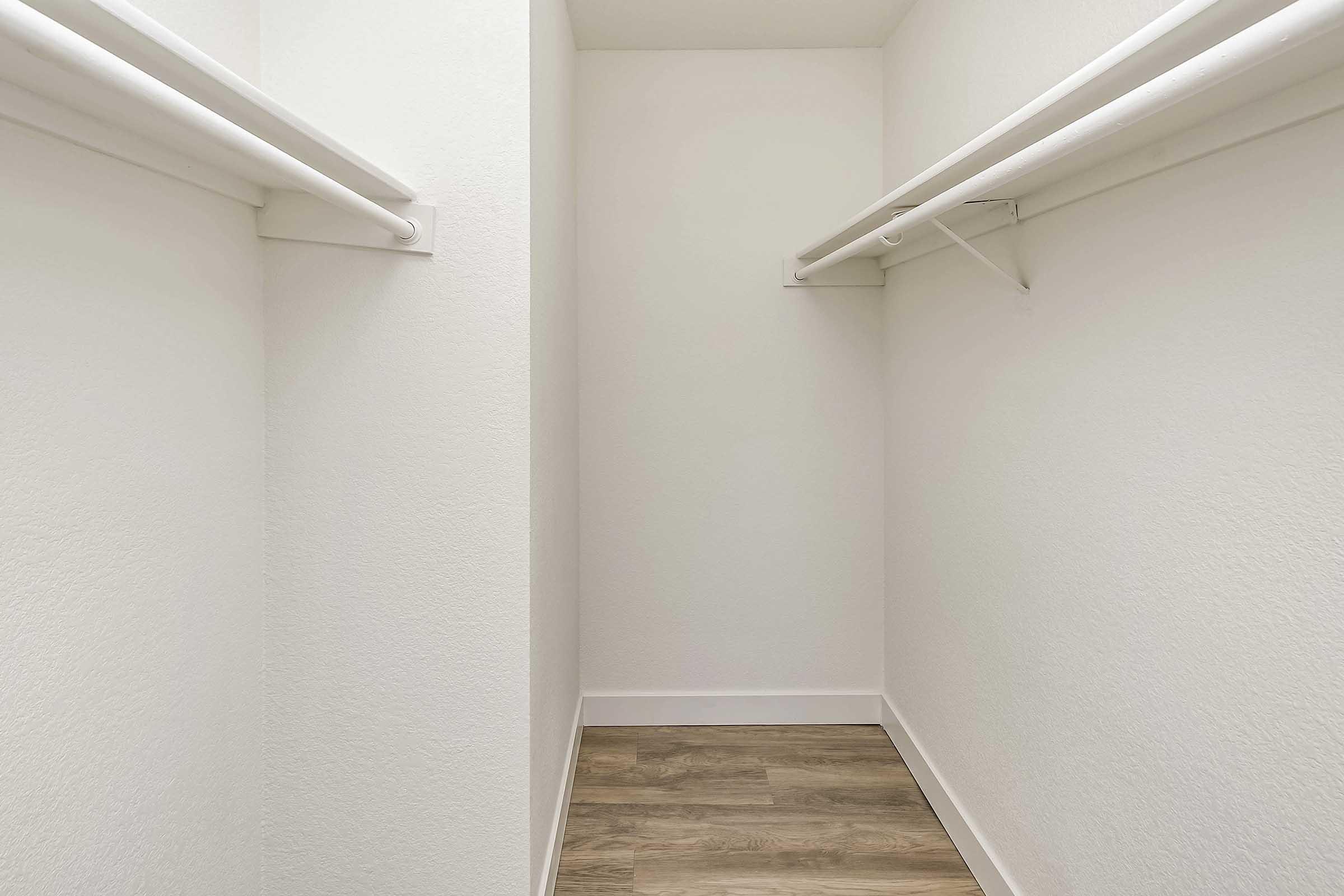 A spacious, empty closet with two white hanging rods mounted on the walls. The walls are light-colored, and the floor features laminate wood flooring, creating a clean and organized look. The closet has no shelves or items, emphasizing its open and minimalist design.
