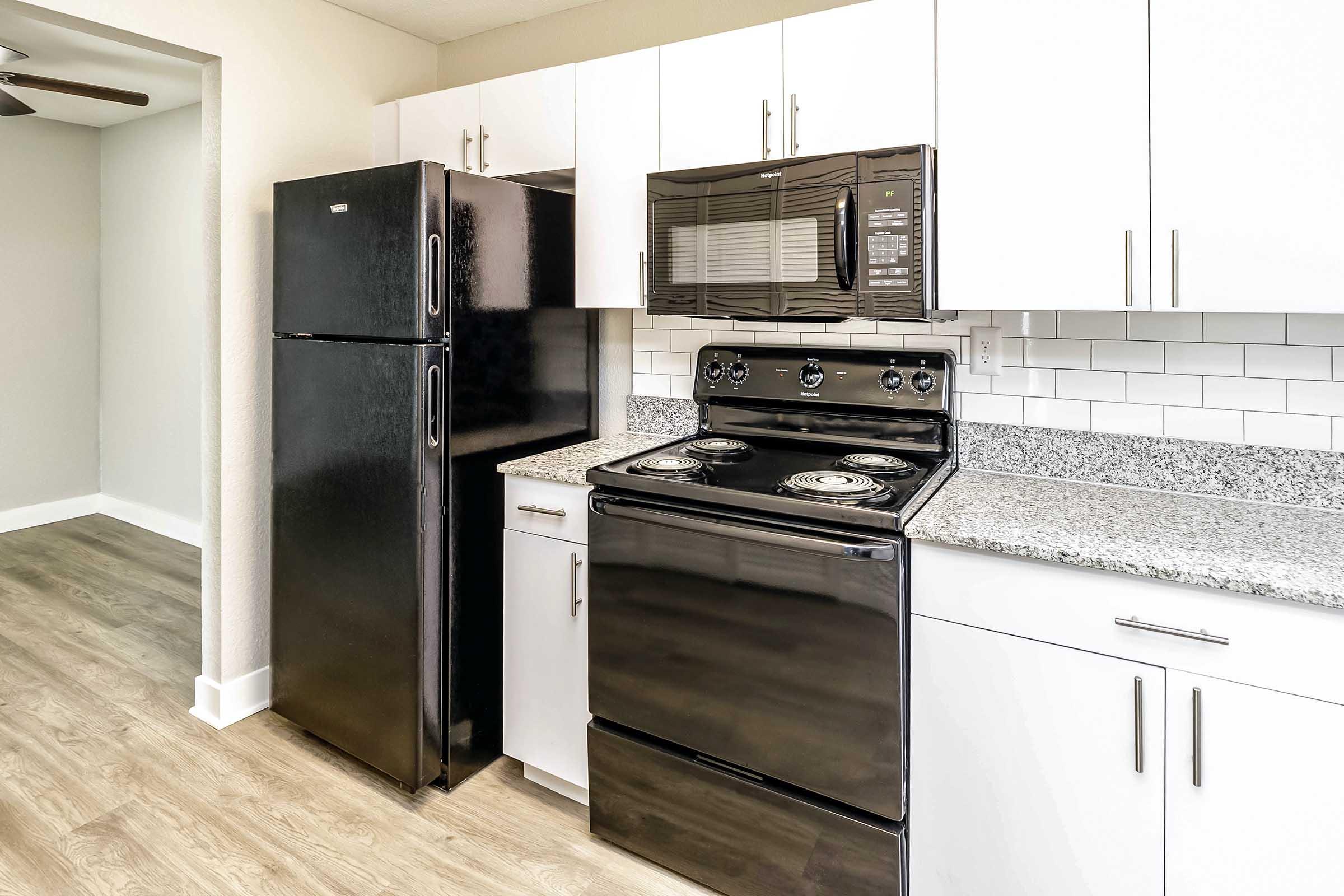 A modern kitchen featuring a black refrigerator, black oven with stovetop, and a sleek microwave above. The cabinetry is white with a granite countertop and a tiled backsplash. The floor is wood-like laminate, and there's a ceiling fan visible in the background.