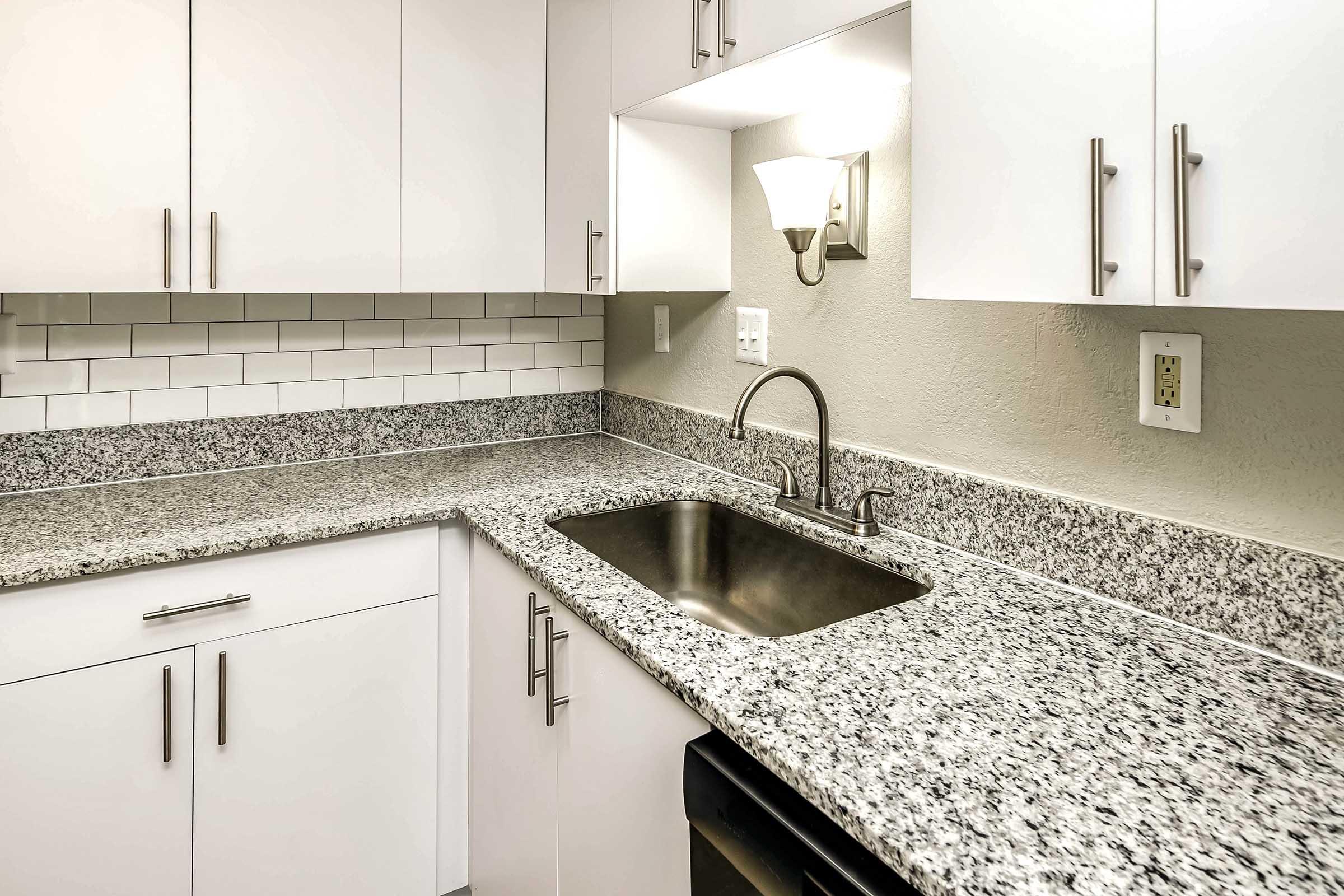 Modern kitchen featuring white cabinetry, a granite countertop, and a stainless steel sink. The backsplash consists of white tiles, and there is ambient lighting from a wall-mounted lamp. A black appliance is partially visible beneath the counter. The overall design is clean and contemporary.