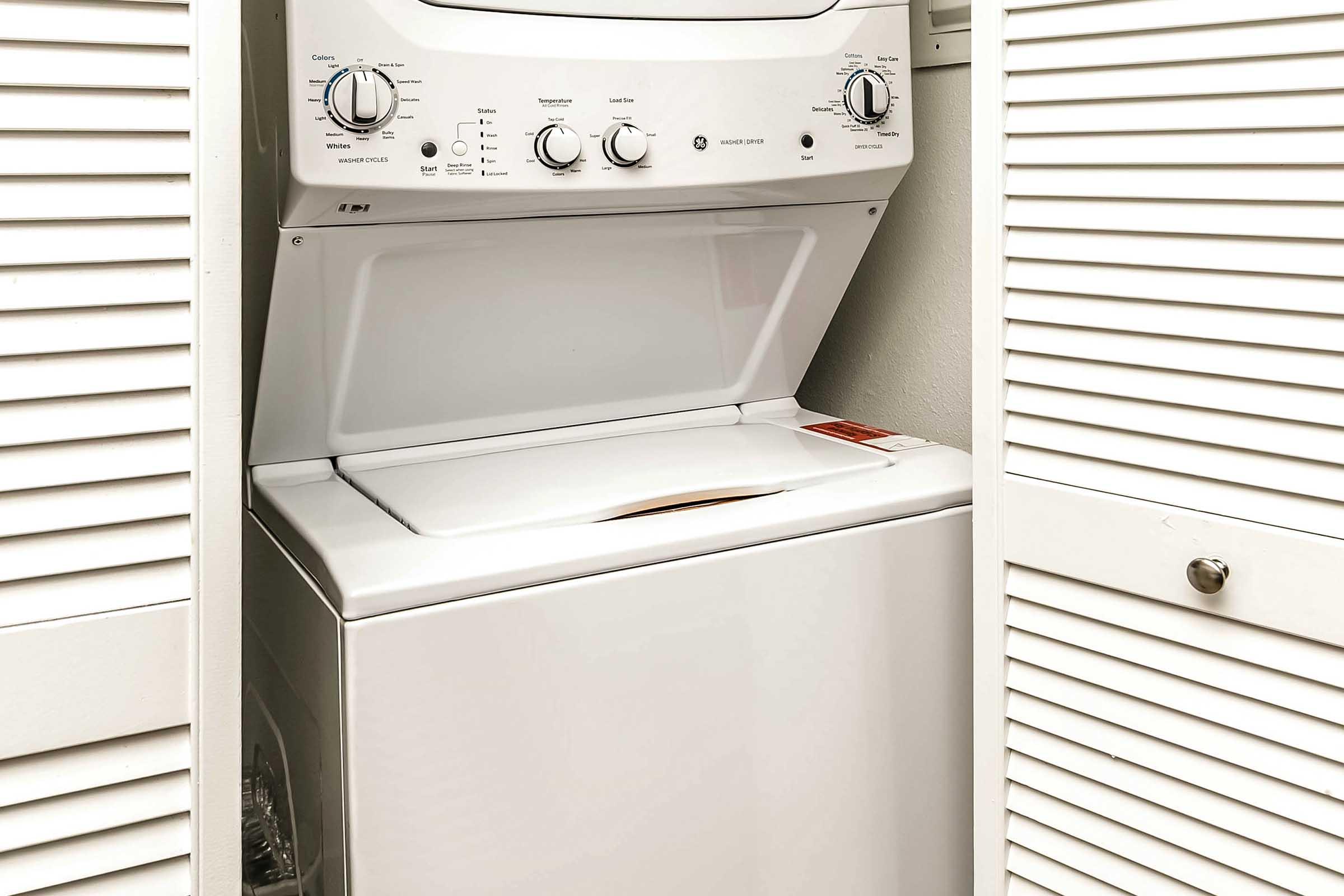 A white stacked washer and dryer unit positioned between two white shuttered doors, showing the controls of the washer on top and the dryer below, in a small laundry space.