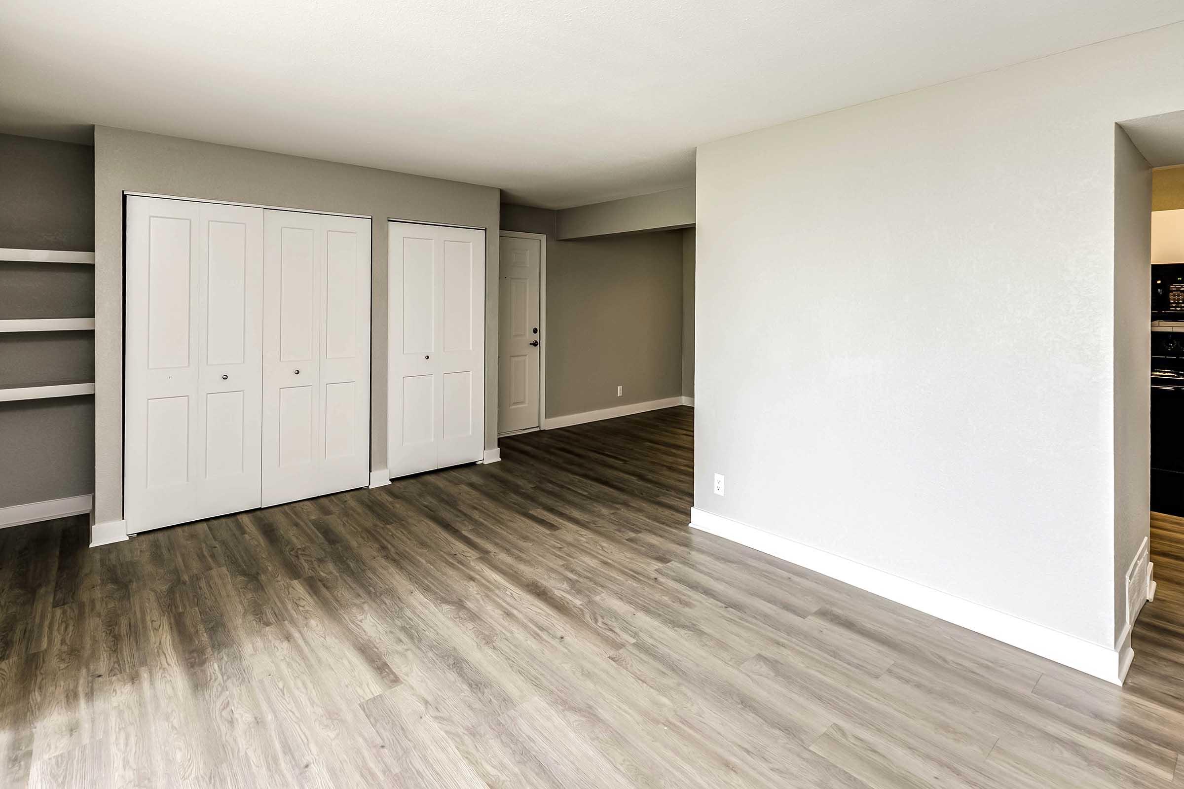 Interior view of a spacious, neutral-toned living area featuring light-colored walls, new laminate flooring, and two white double closets. There is an adjacent wall with an opening leading to a kitchen area. The room has a bright, airy feel with ample natural light.