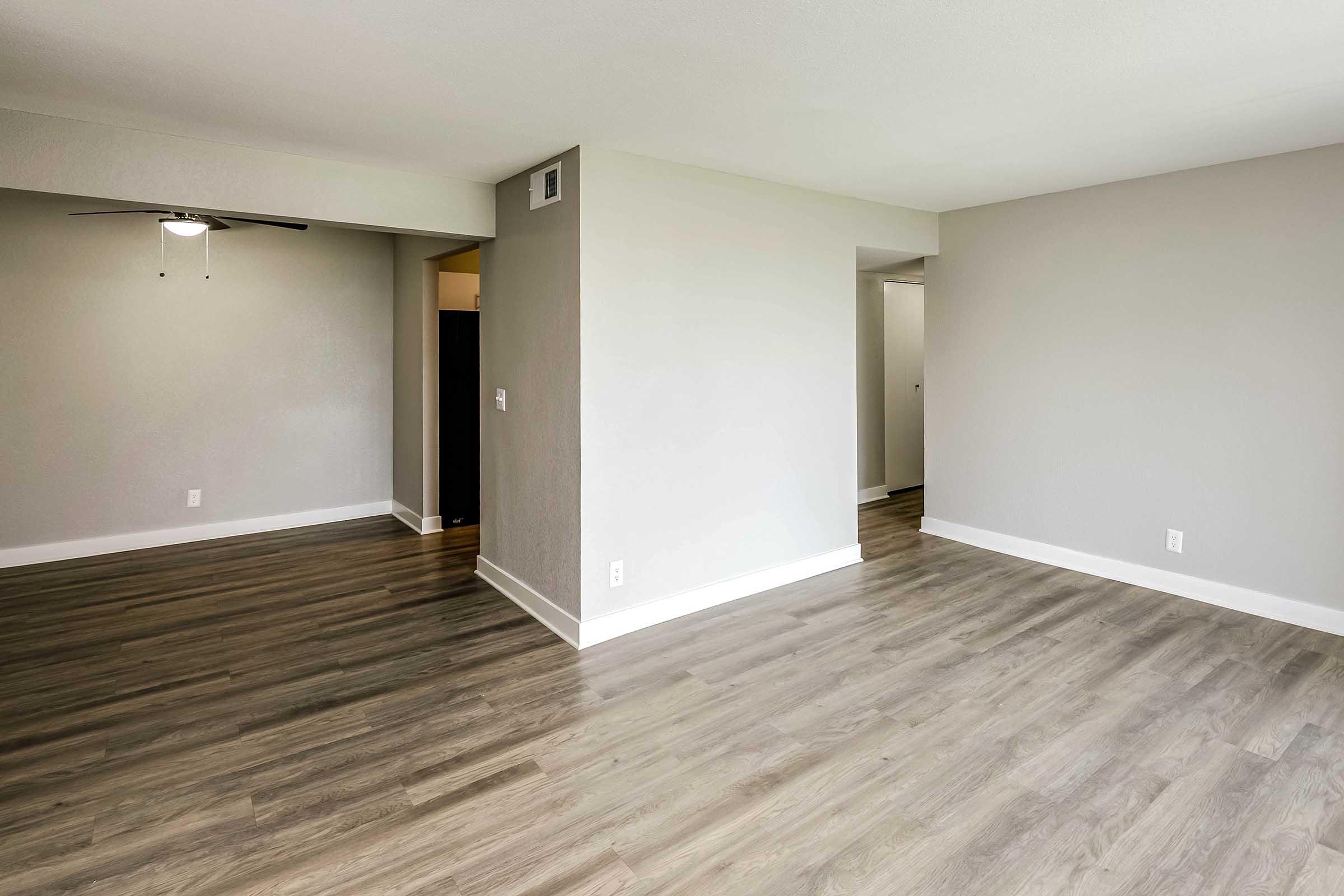 A spacious, empty room with light gray walls and a wood-like floor. There is a ceiling fan visible on the left, and the room features an open layout leading to a doorway. Natural light brightens the space, highlighting its clean lines and modern design.