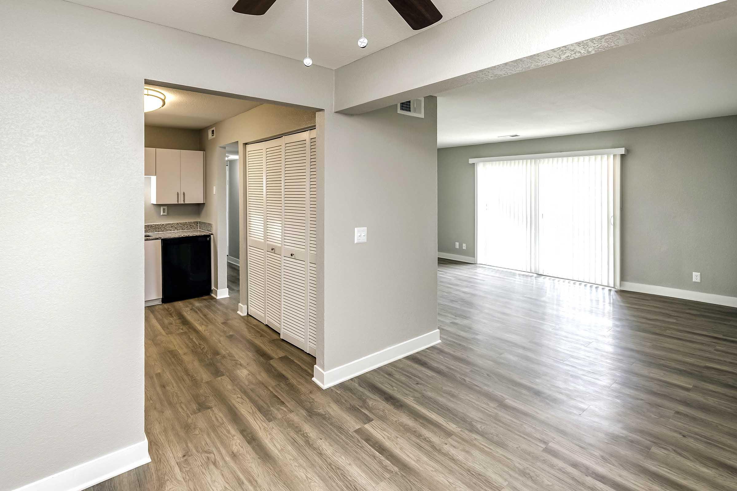 Interior view of a modern living space featuring a light gray wall color, laminate flooring, and a ceiling fan. On the left, there is a doorway leading to a kitchen with cabinets, and on the right, large sliding glass doors allow natural light into the room, which is open and spacious.