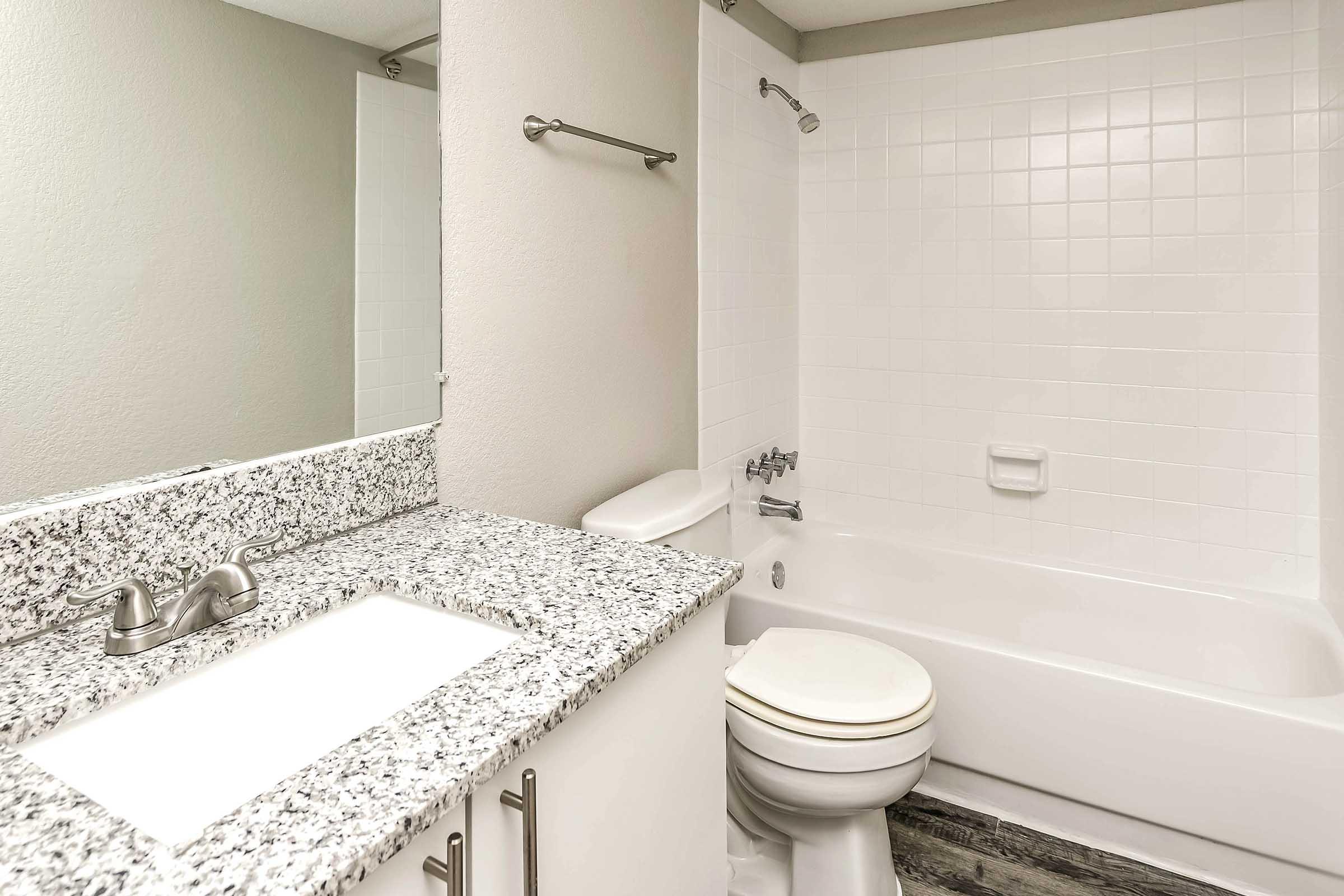 Bright and modern bathroom featuring a granite countertop sink with a mirror above, a white bathtub with a showerhead, and light-colored tiled walls. The floor has a wood-like finish, adding warmth to the space.