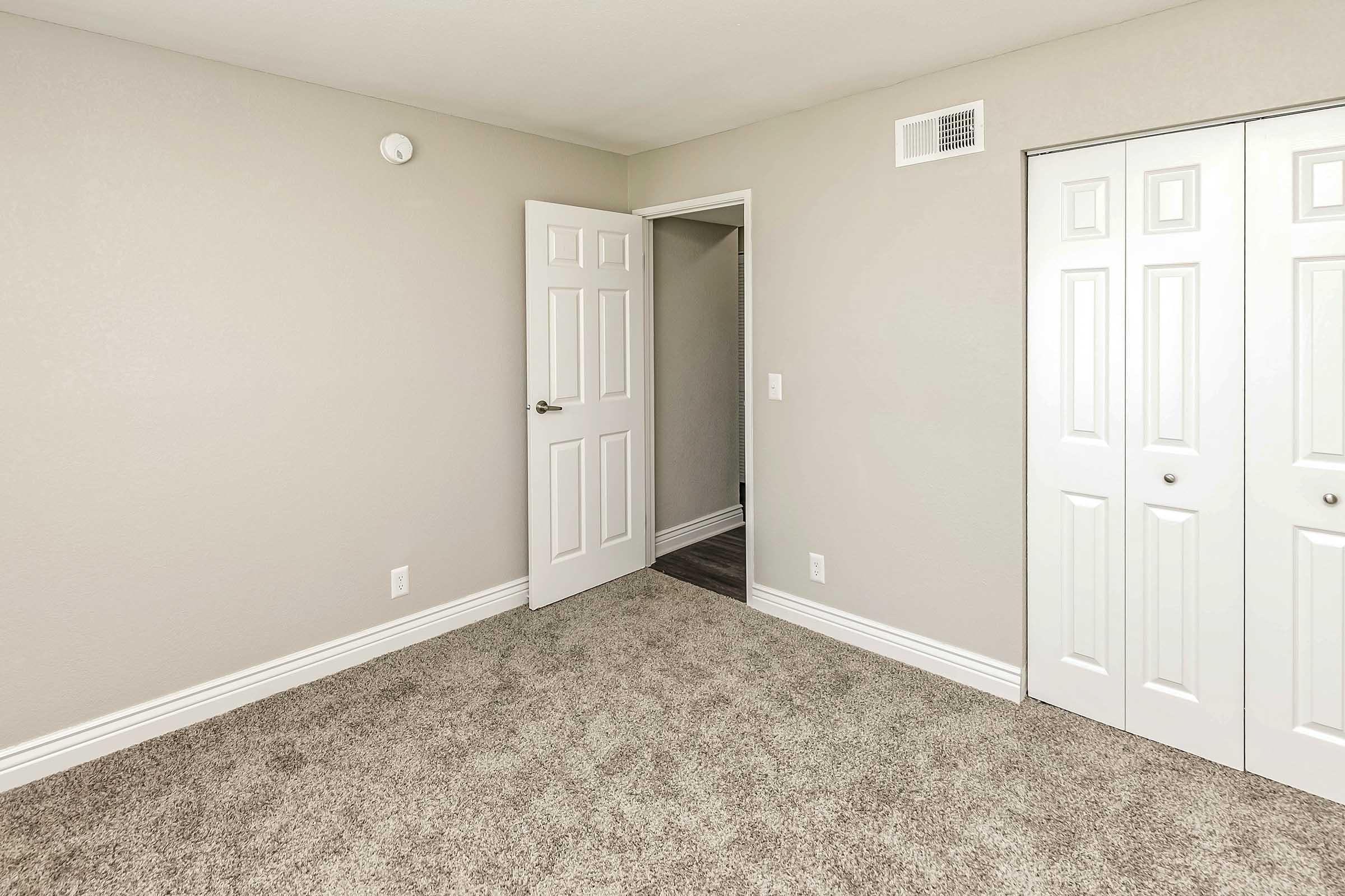 A clean, empty room with freshly painted light gray walls and beige carpet. A white door is ajar, leading to another room, while a closet with double white doors is visible on the right. Natural light brightens the space, creating a welcoming atmosphere.