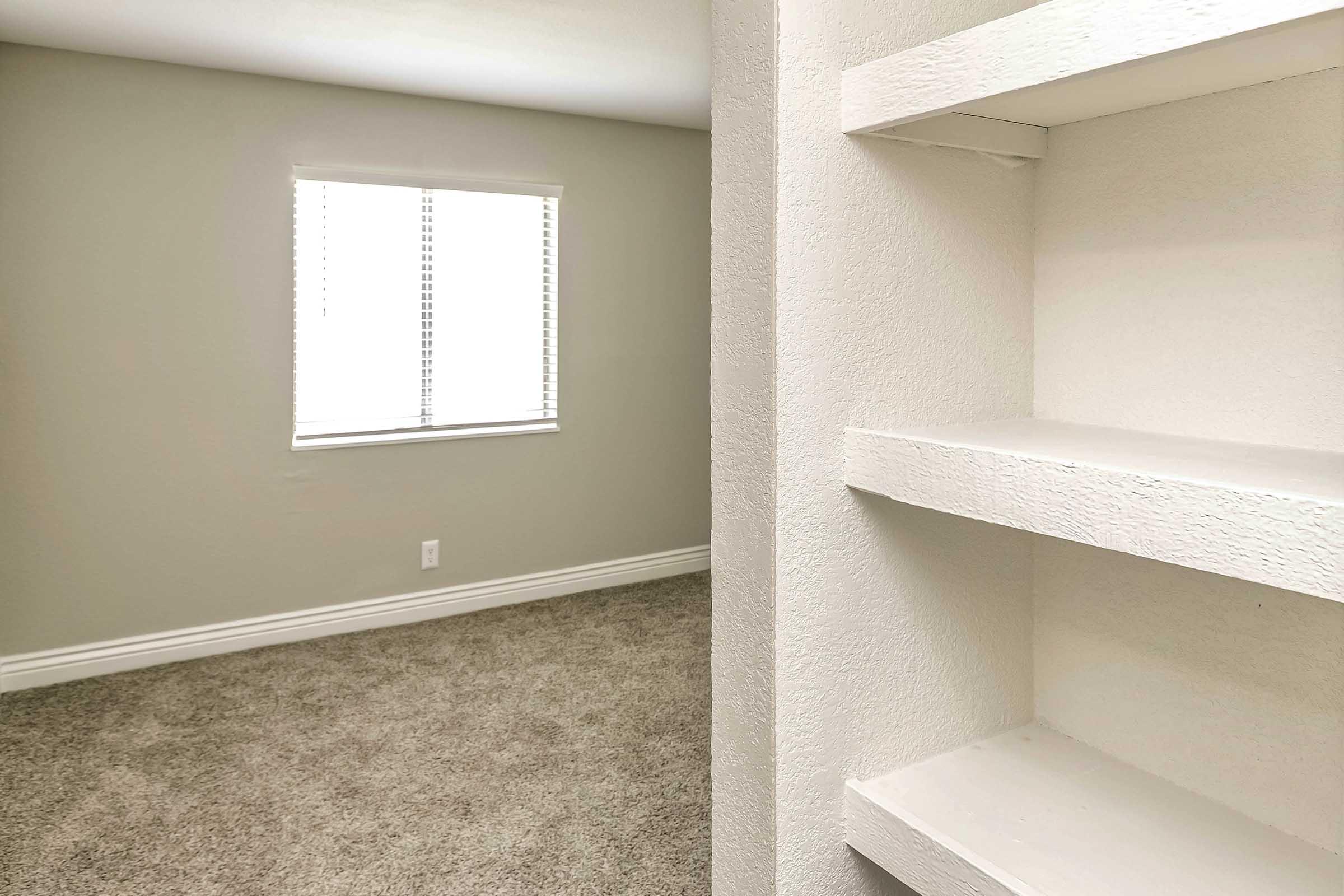 A spacious, well-lit room featuring a window with blinds, beige walls, and plush carpeting. On the right, there is a close-up view of a white shelf against the wall, suggesting a clean and modern design. The overall atmosphere is bright and inviting, perfect for various uses.