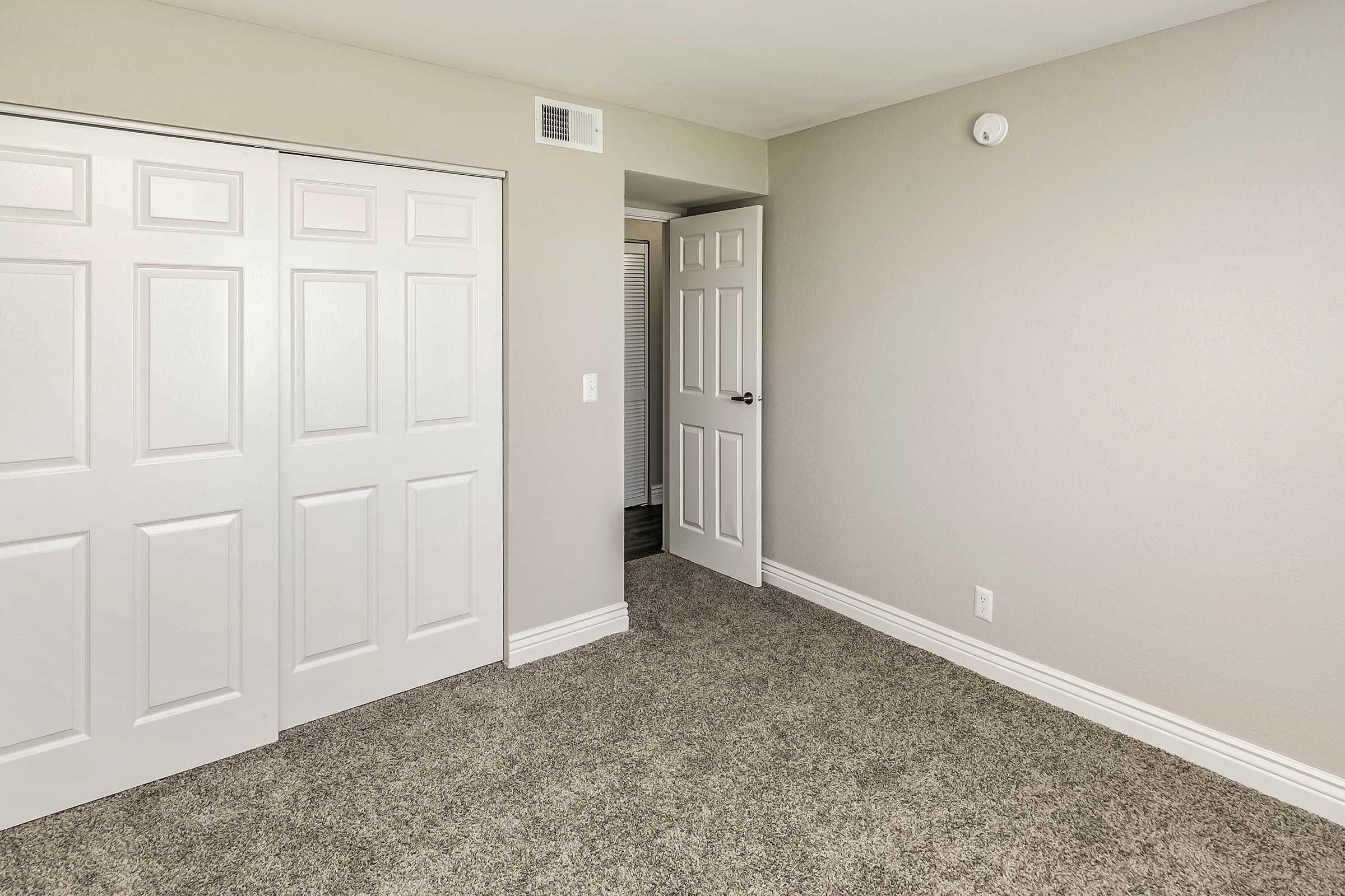 A well-lit room with beige walls and a plush carpet. There are double white closet doors on one side and a single door leading to another space. The room is empty and minimalistic, featuring a neutral color palette and modern design elements.