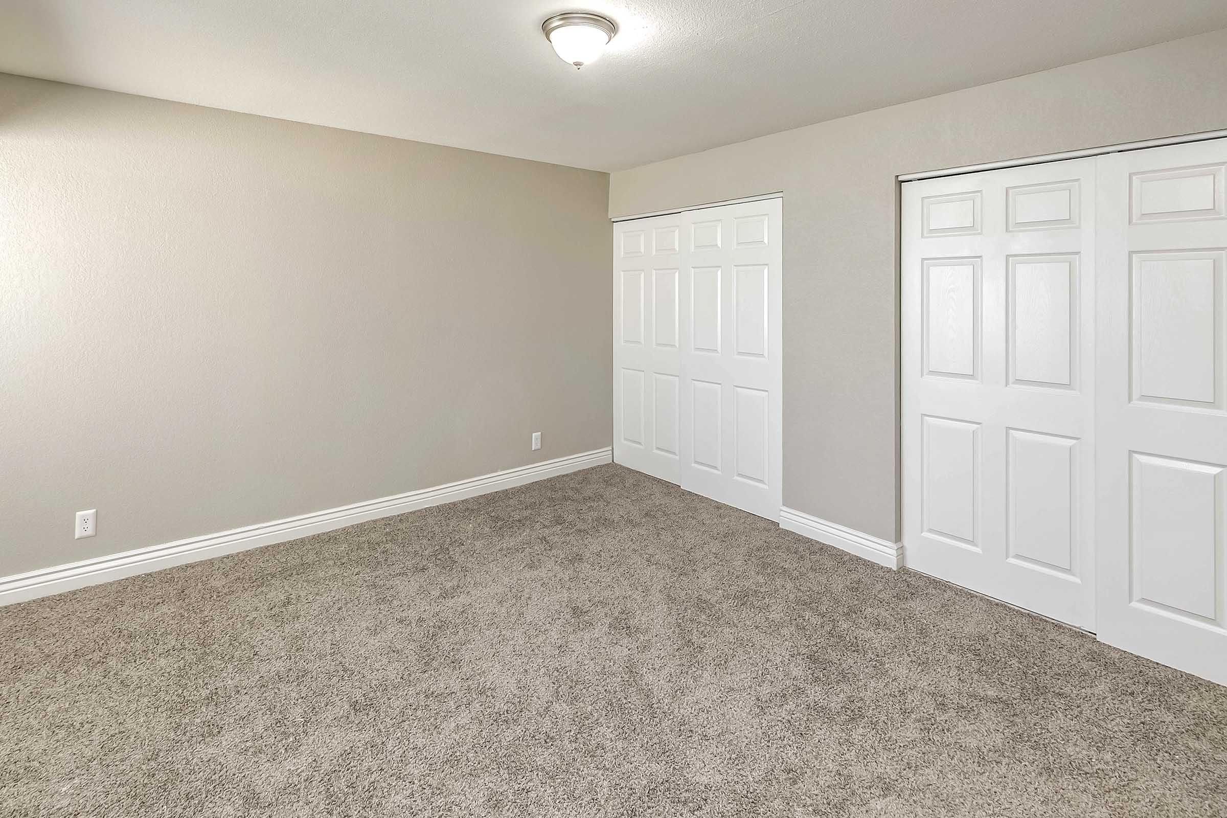 Empty room with light beige walls and carpet, featuring a ceiling light fixture. Two white sliding closet doors are visible on one wall, and the space is well-lit and minimalistic, creating an inviting atmosphere. No furniture is present.