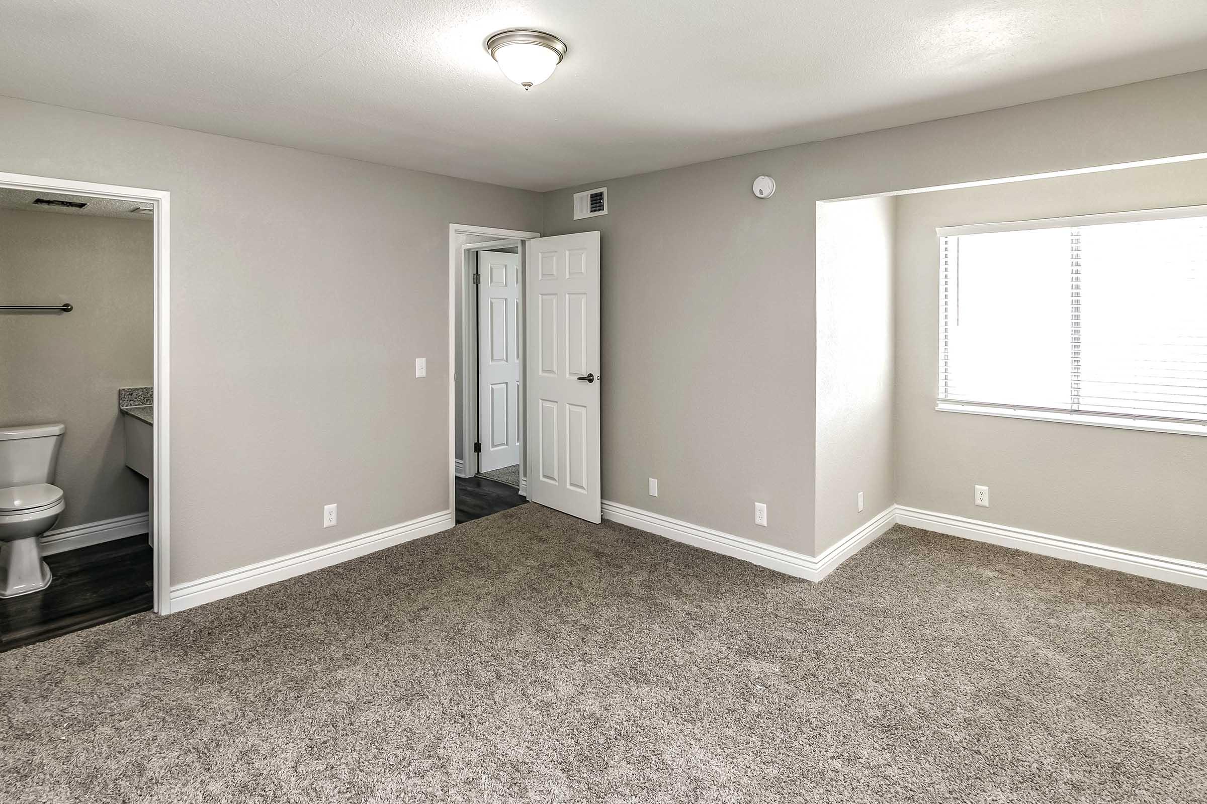 A spacious, light-colored room with beige carpeting and white walls. Two doorways lead to adjacent spaces, one with a toilet visible. A window allows natural light, and a ceiling light fixture is visible. The room is empty, creating a clean and fresh atmosphere.