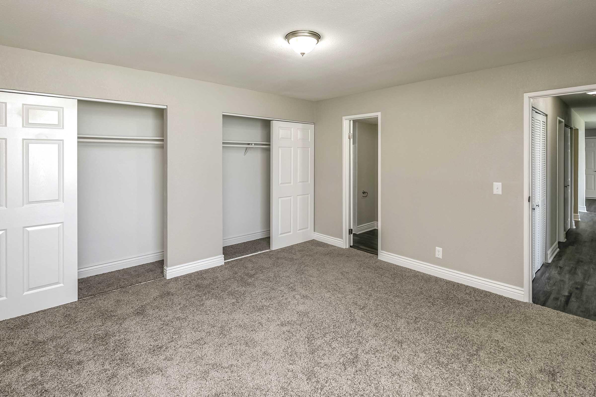 Empty room with light gray walls, a beige carpet, and a ceiling light fixture. Two closets with white sliding doors are on one wall, and a door leads to another room on the opposite side. The space appears spacious and well-lit, suitable for various purposes.