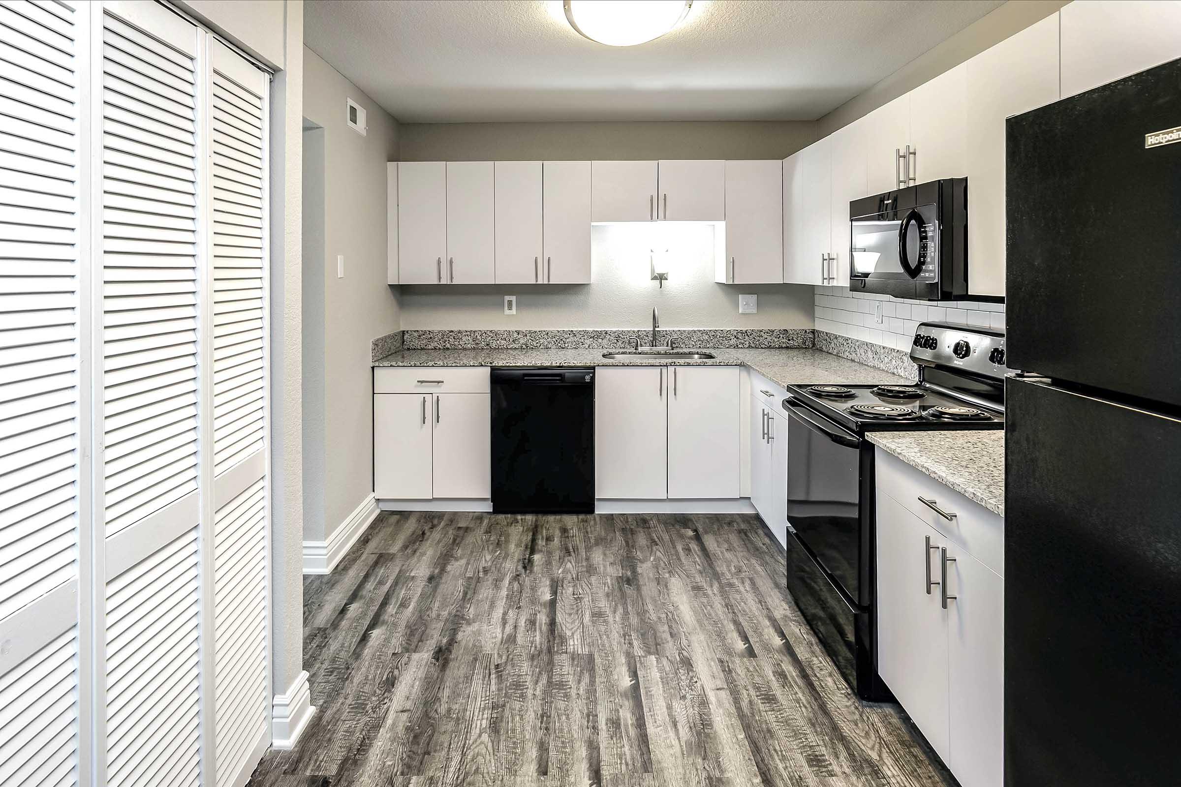 Modern kitchen featuring white cabinetry, granite countertops, stainless steel appliances including a microwave and stove, a black refrigerator, and dark wood-look flooring. The space is well-lit with an overhead light, and the layout is open with a slight angle towards the entryway.