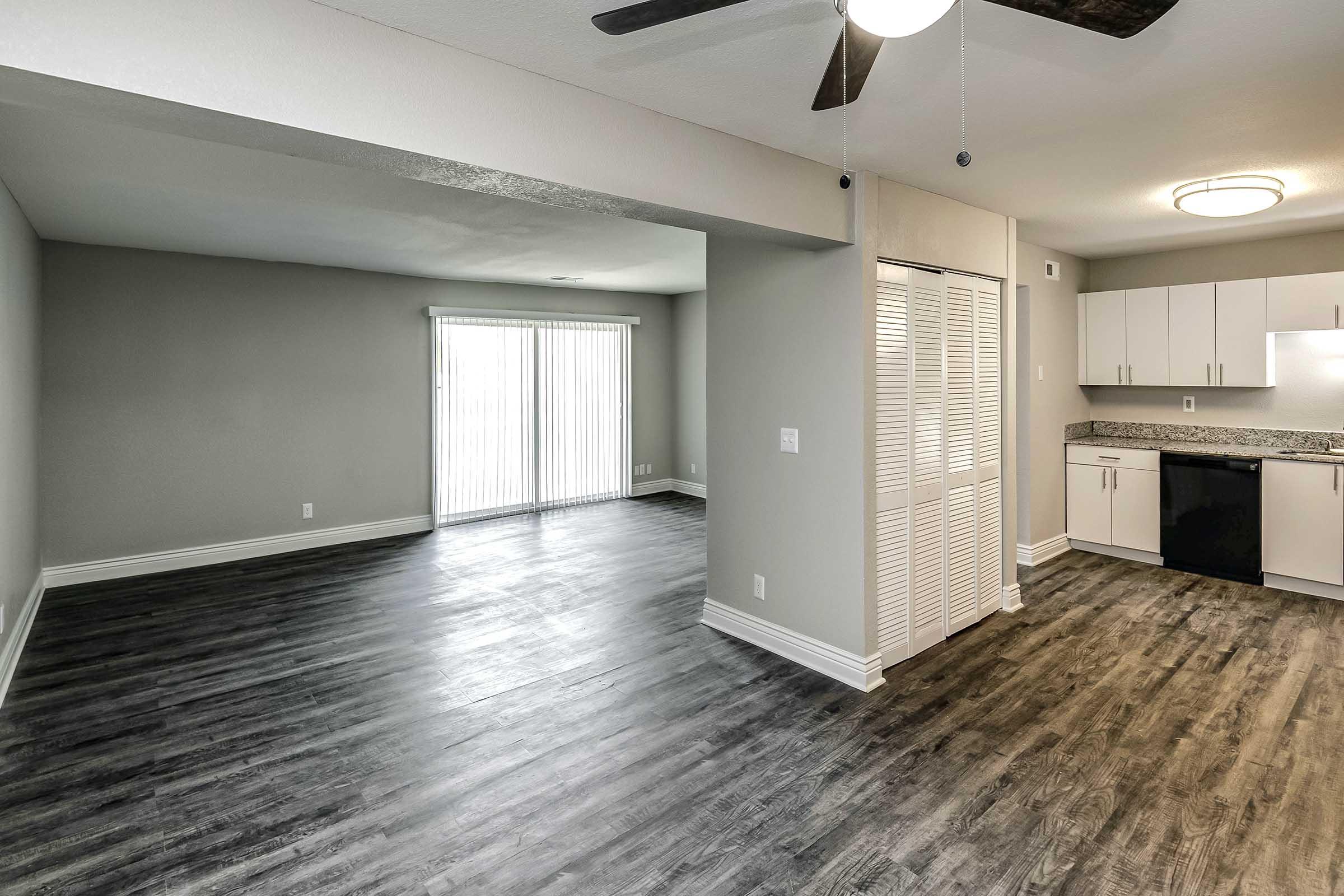 A modern, open-concept living space featuring a living area with large sliding glass doors leading to the outside, and a kitchen area equipped with white cabinets and dark countertops. The flooring is dark wood laminate, providing a contemporary look. Ceiling fan installed for ventilation.