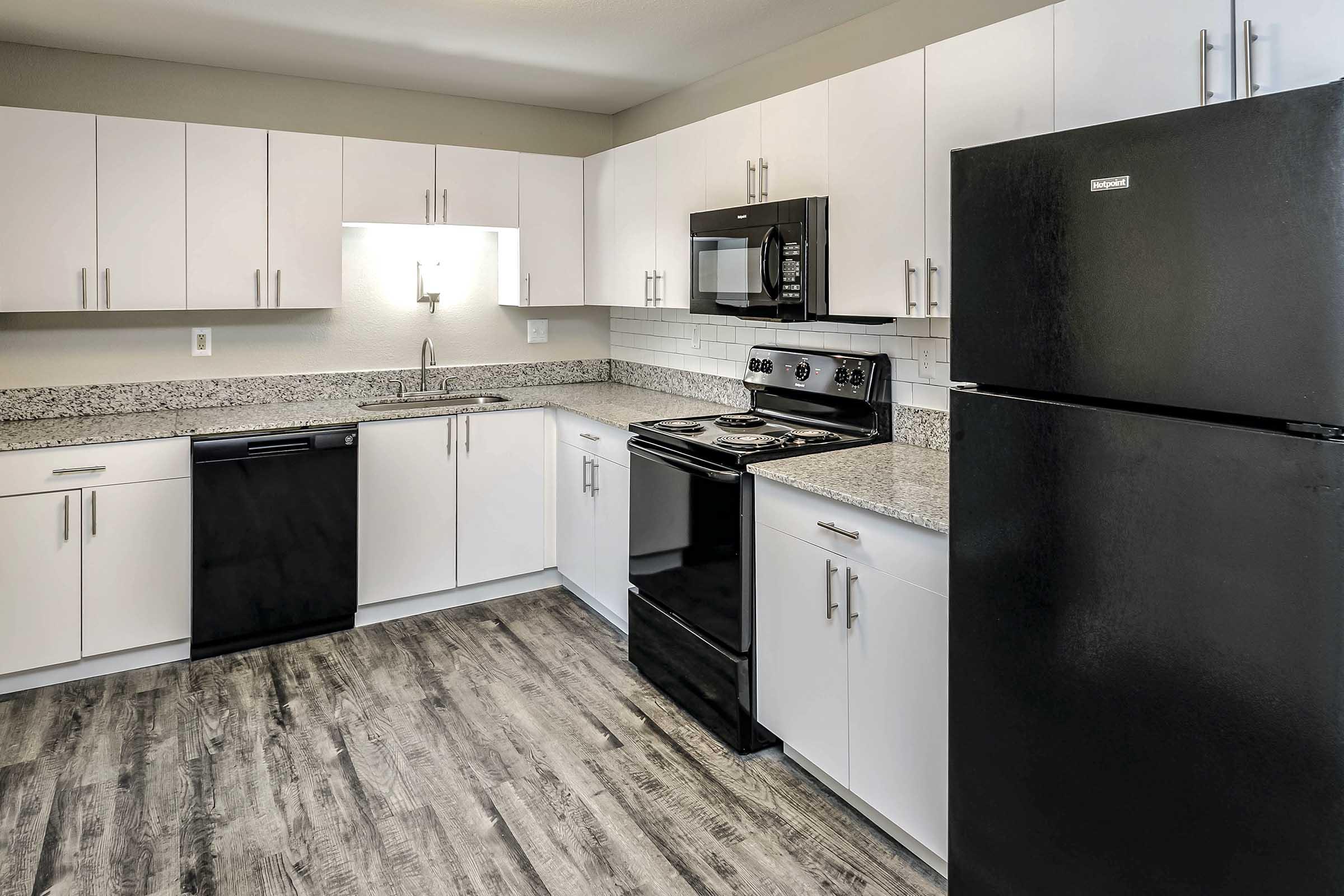 Modern kitchen featuring white cabinets, gray granite countertops, stainless steel sink, black appliances including a refrigerator, microwave, and stove, along with a dishwasher. The floor is a wood-like laminate, creating a stylish and functional cooking space.