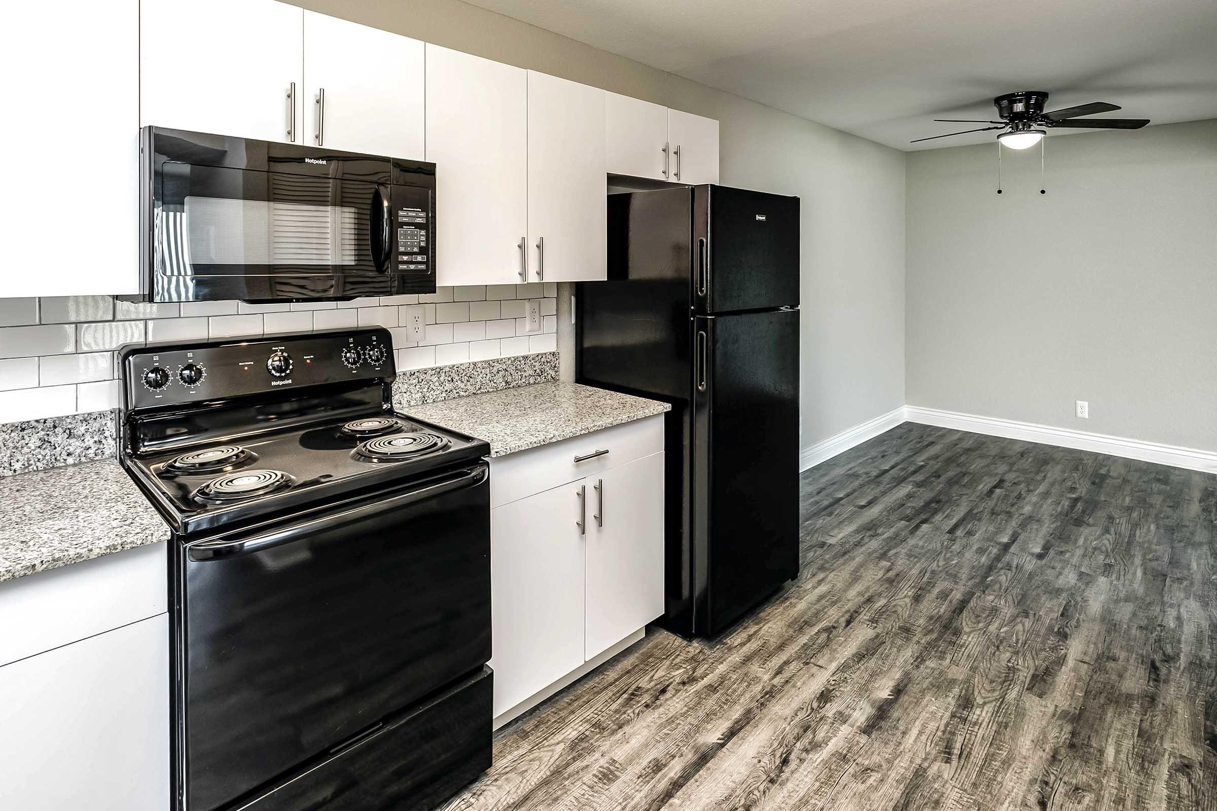 Modern kitchen featuring black appliances, including an electric stove and a refrigerator, set against white cabinets and a granite countertop. The flooring is a sleek, dark wood laminate, and the space is well-lit with a neutral wall color and a ceiling fan visible in the adjoining area.
