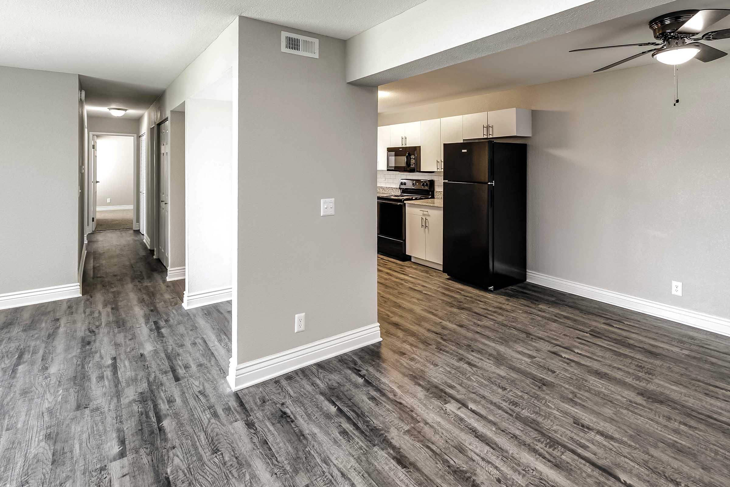 Interior view of a modern apartment featuring a spacious, open layout. The room has light gray walls, dark wood-like flooring, and a ceiling fan. A kitchen is visible at the end with black appliances, including a refrigerator and stove. A hallway leads to additional rooms. Natural light comes from ceiling fixtures.