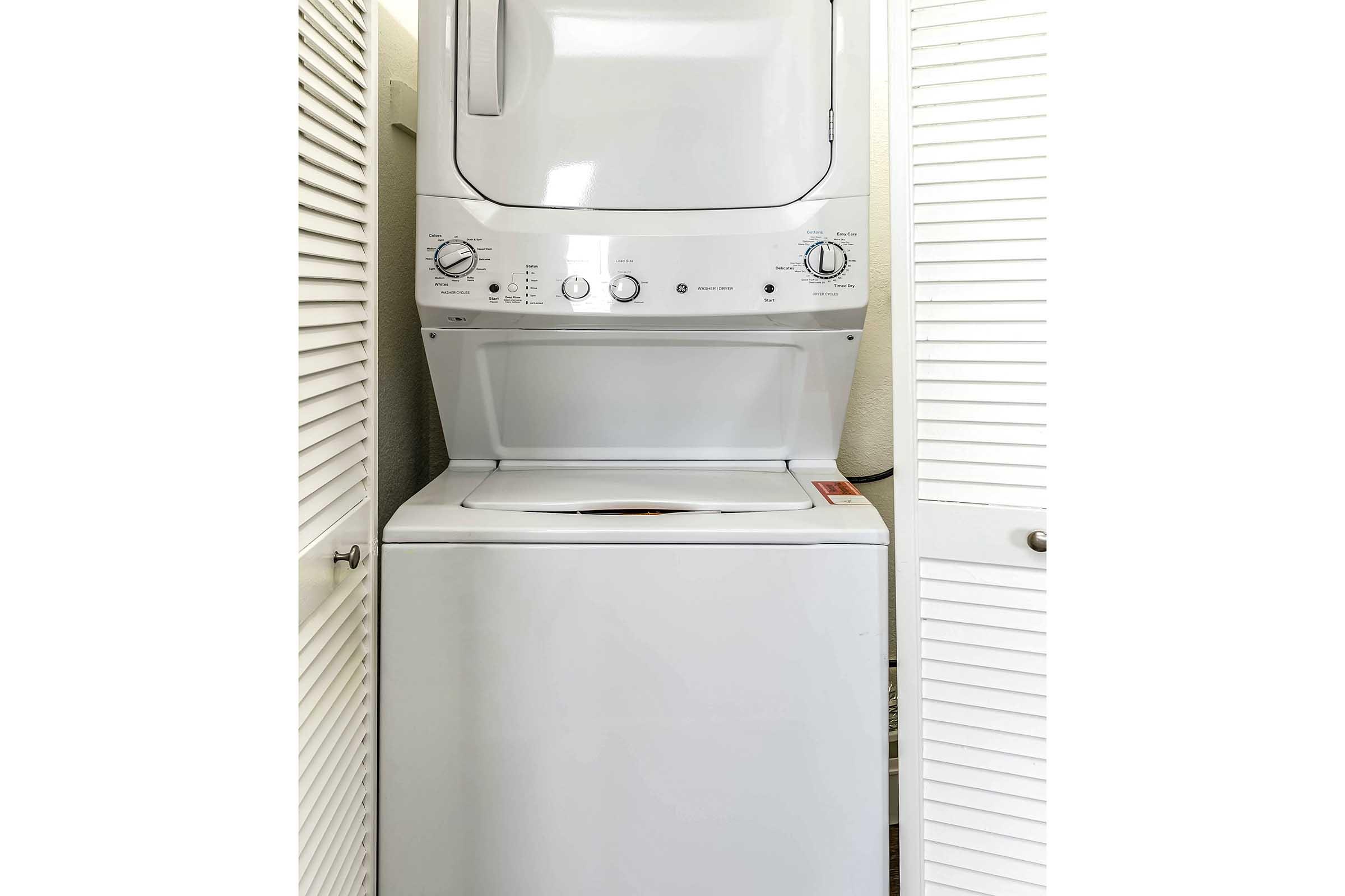 A stacked washer and dryer unit located in a compact laundry space, framed by closed bifold doors. The appliance features control knobs and settings for various washing and drying cycles. The unit is modern and white, suitable for small areas.