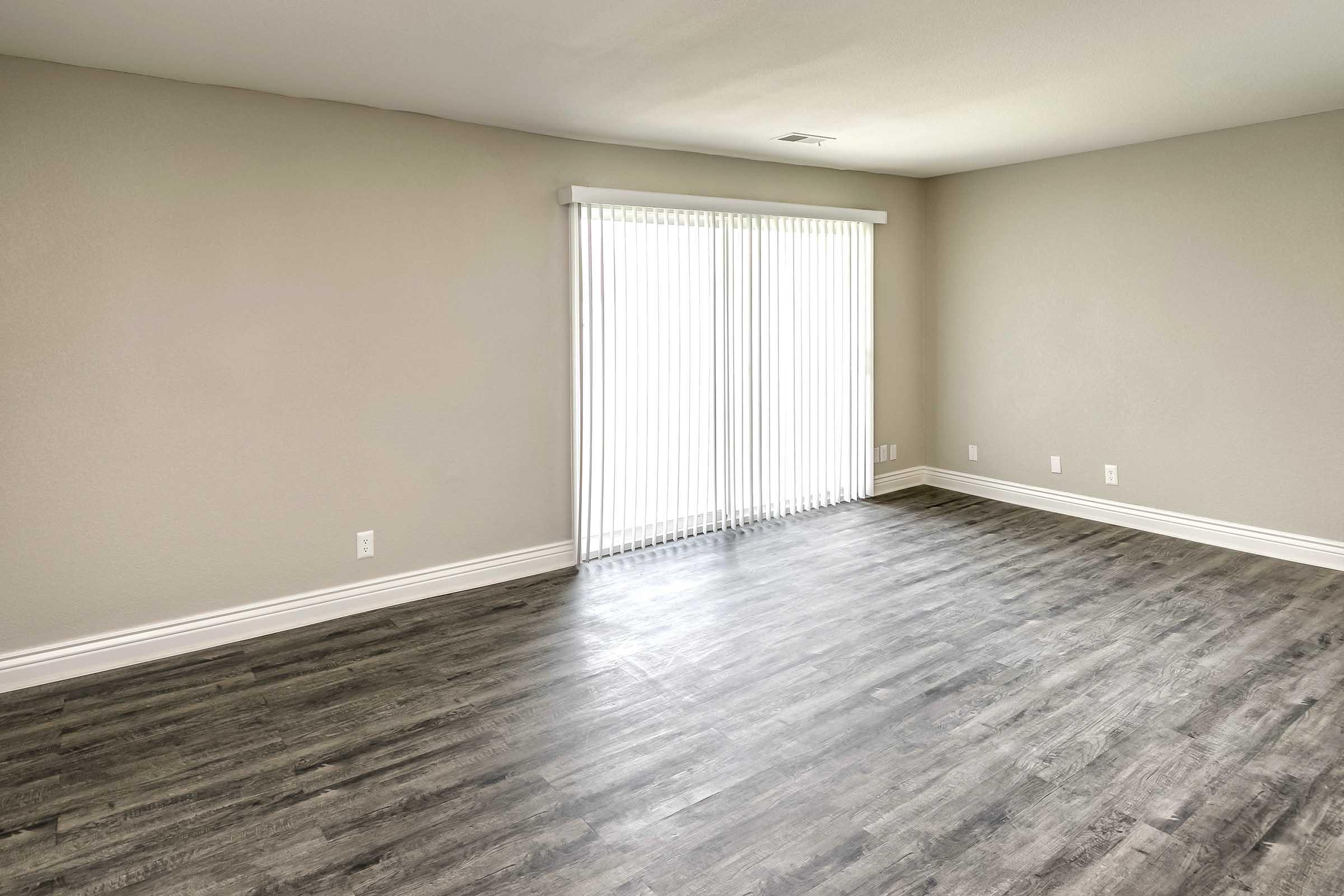 Empty room with light gray walls and new wood-like flooring. A large window with vertical blinds allows natural light to fill the space, creating a bright and airy atmosphere. There are no furnishings, showcasing the simplicity and openness of the area.