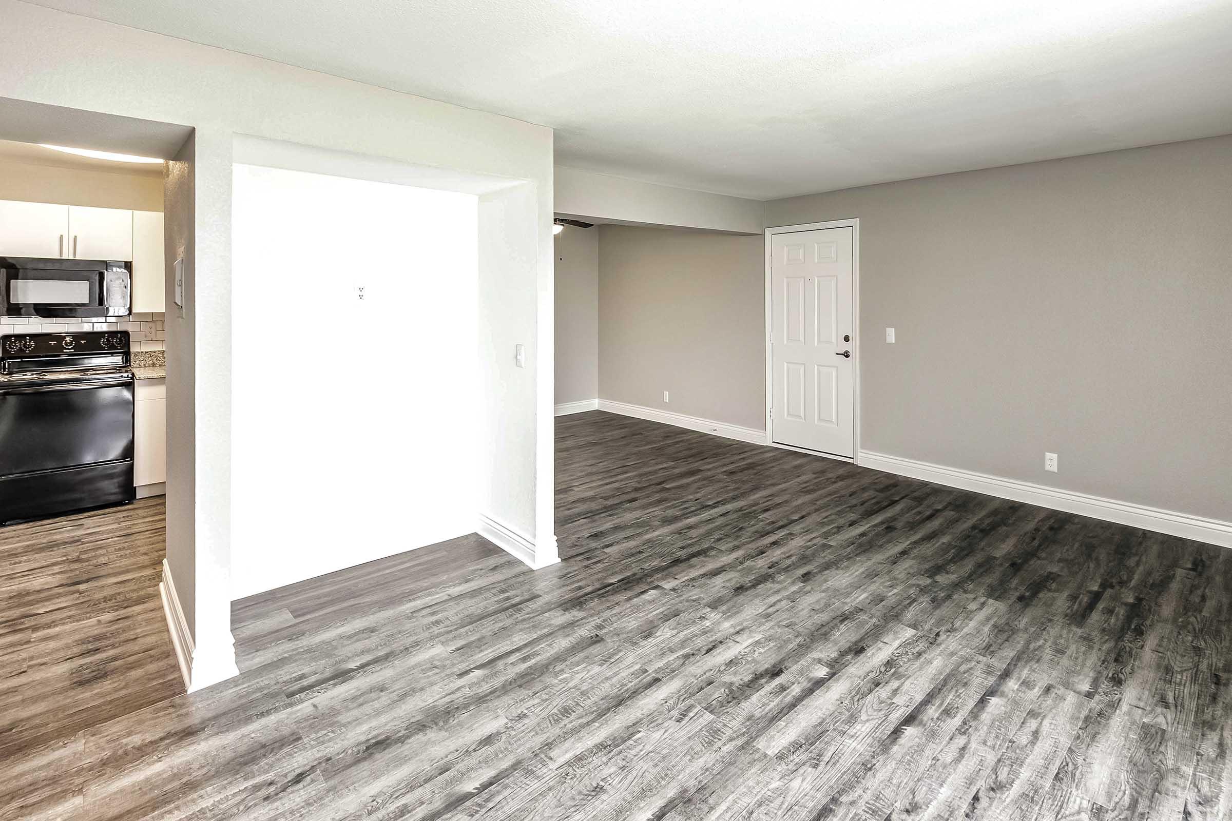 A spacious, empty room featuring light gray walls and a wood-look floor. On the left, there’s a modern kitchen with dark appliances, and on the right, a white door leading to another space. The room has large windows allowing natural light to enter.