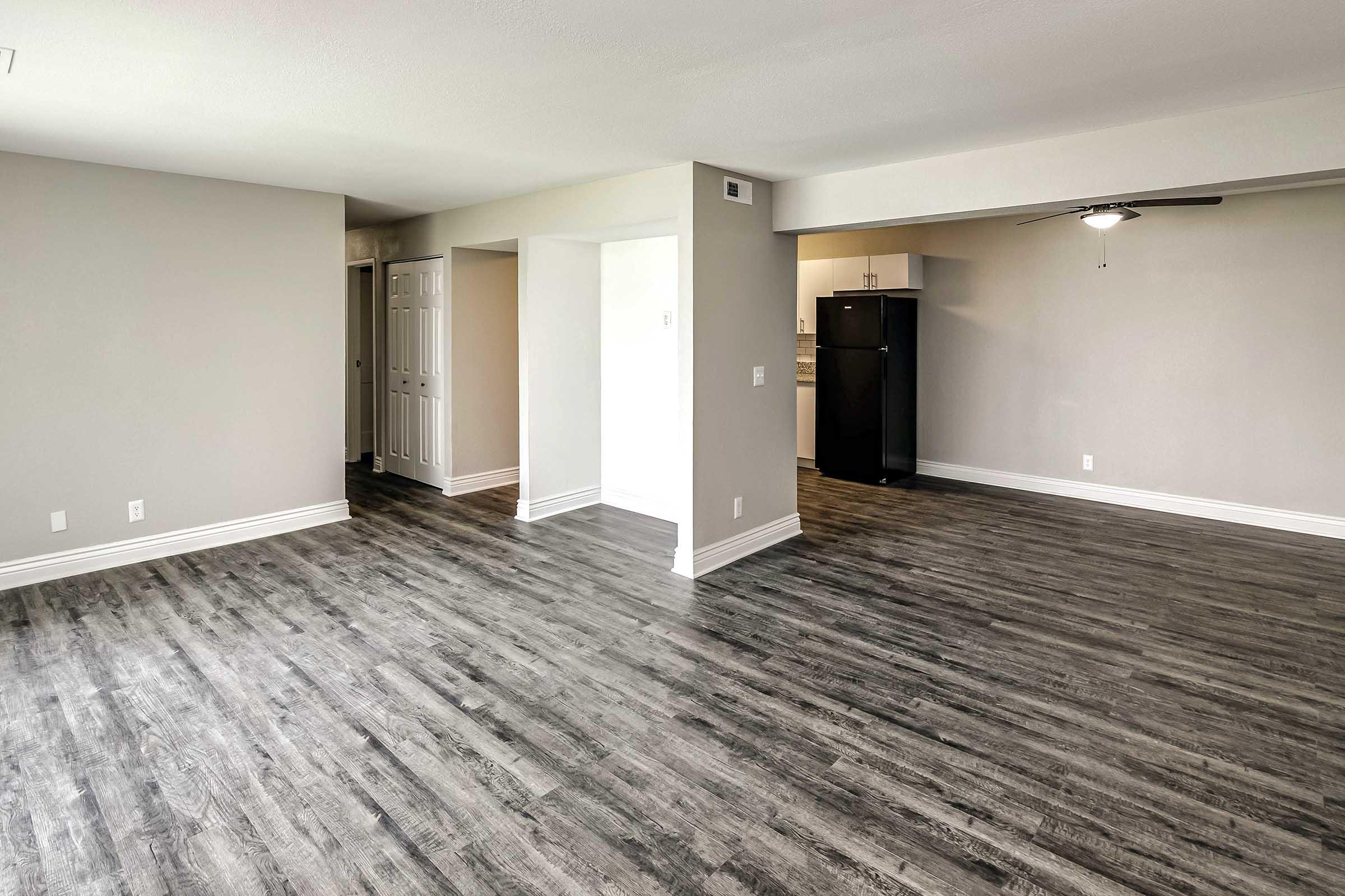 A spacious, modern living area featuring light gray walls and dark wood-like flooring. The room has a ceiling fan, an open layout leading to a kitchenette with a black refrigerator. Natural light brightens the space, making it inviting and ready for furnishings.