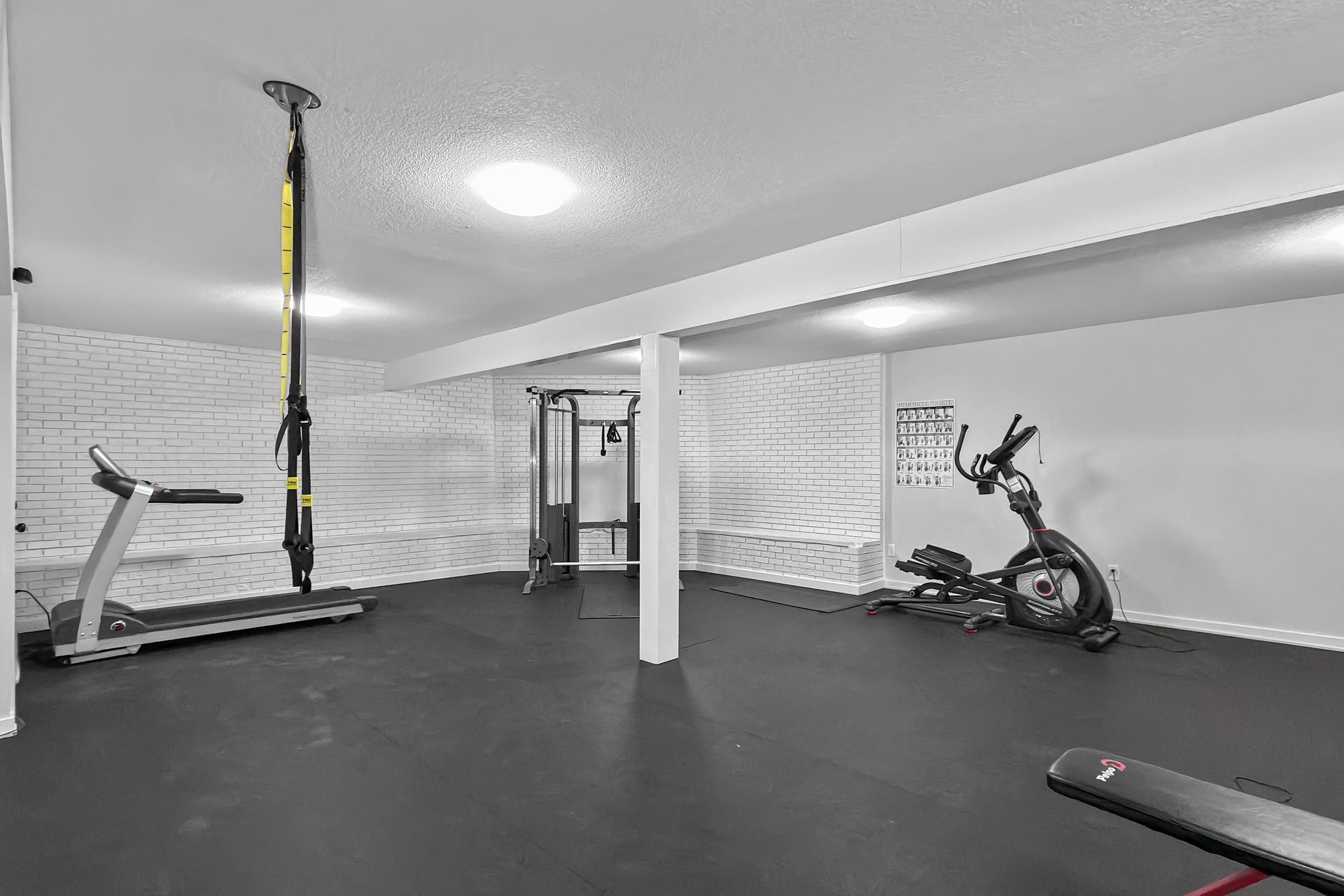A spacious gym interior featuring various exercise equipment. There's a treadmill, an elliptical machine, and a cable machine. Suspension training straps hang from the ceiling. The walls are white brick, and the flooring is dark. Bright overhead lighting illuminates the area, creating a clean and modern workout space.