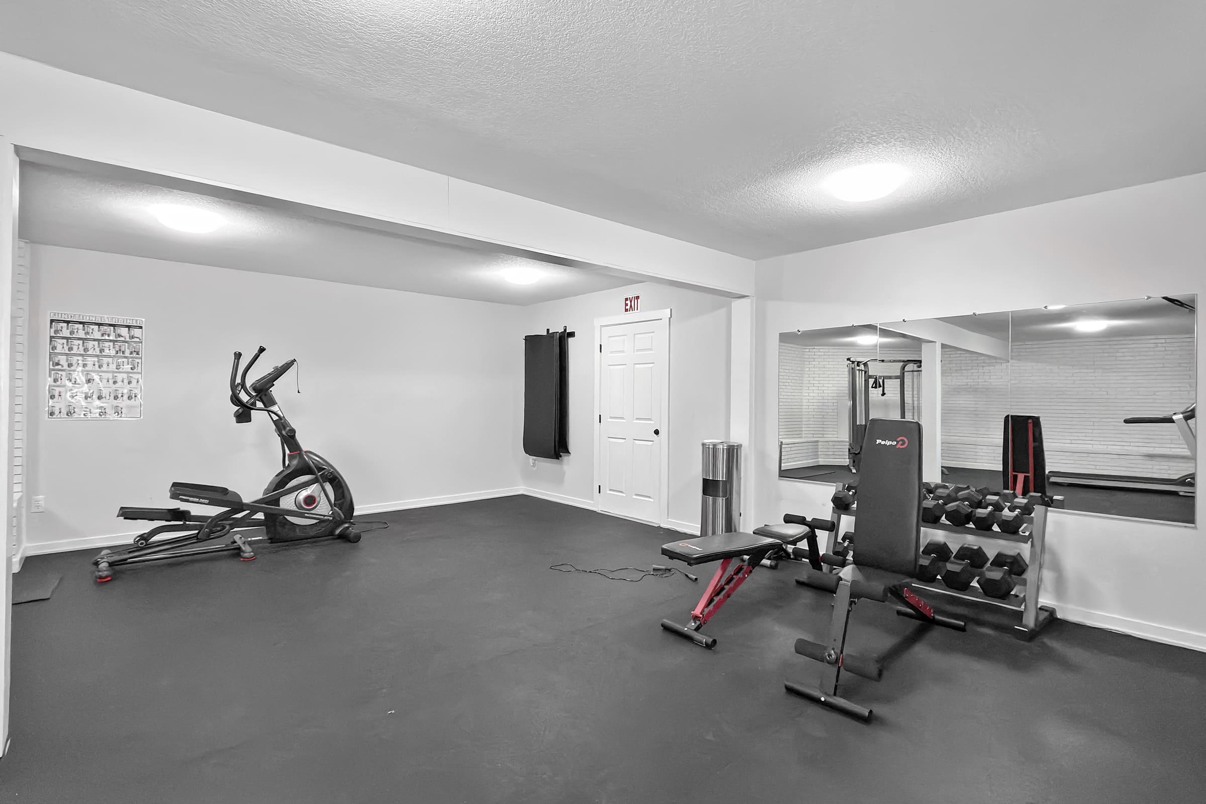 A modern home gym featuring an elliptical machine, a weight bench with dumbbells, a mirror on one wall, and a black mat flooring. There is a wall-mounted black punching bag and a door leading to another room. Adequate lighting creates a bright and spacious atmosphere.