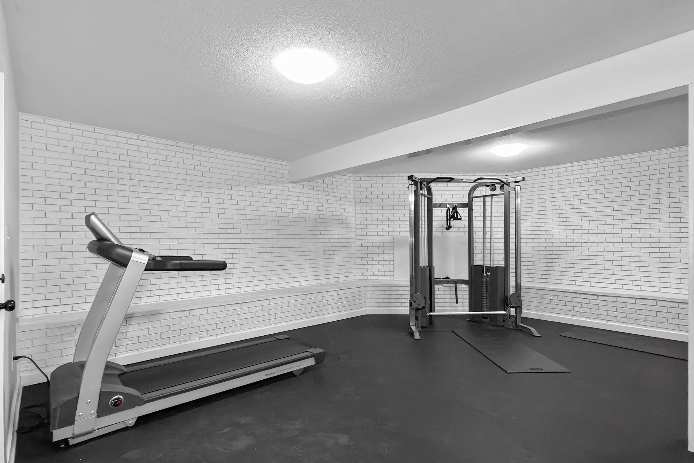 A spacious home gym featuring a treadmill and a multi-station strength training machine. The walls are white brick, and the floor is dark, creating a modern and clean workout environment with ample natural light.