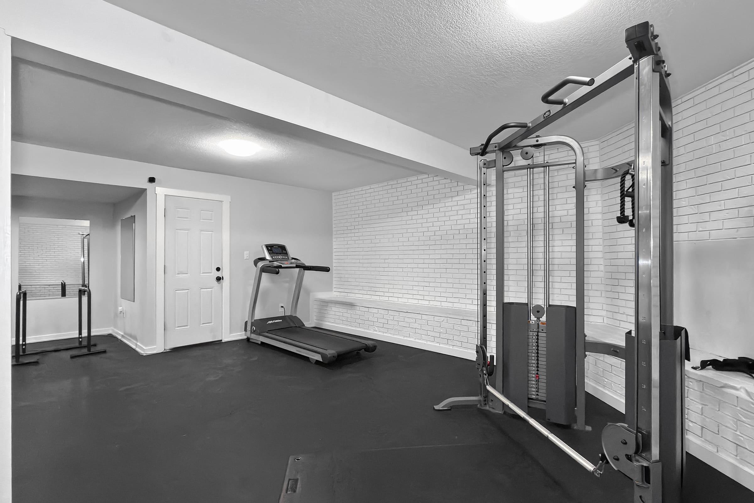 A clean and minimalist home gym with a cable machine, treadmill, and mirrored wall. The floor is dark, and the walls are white, creating a spacious and bright environment for workouts. A door is visible in the background, leading to another room.