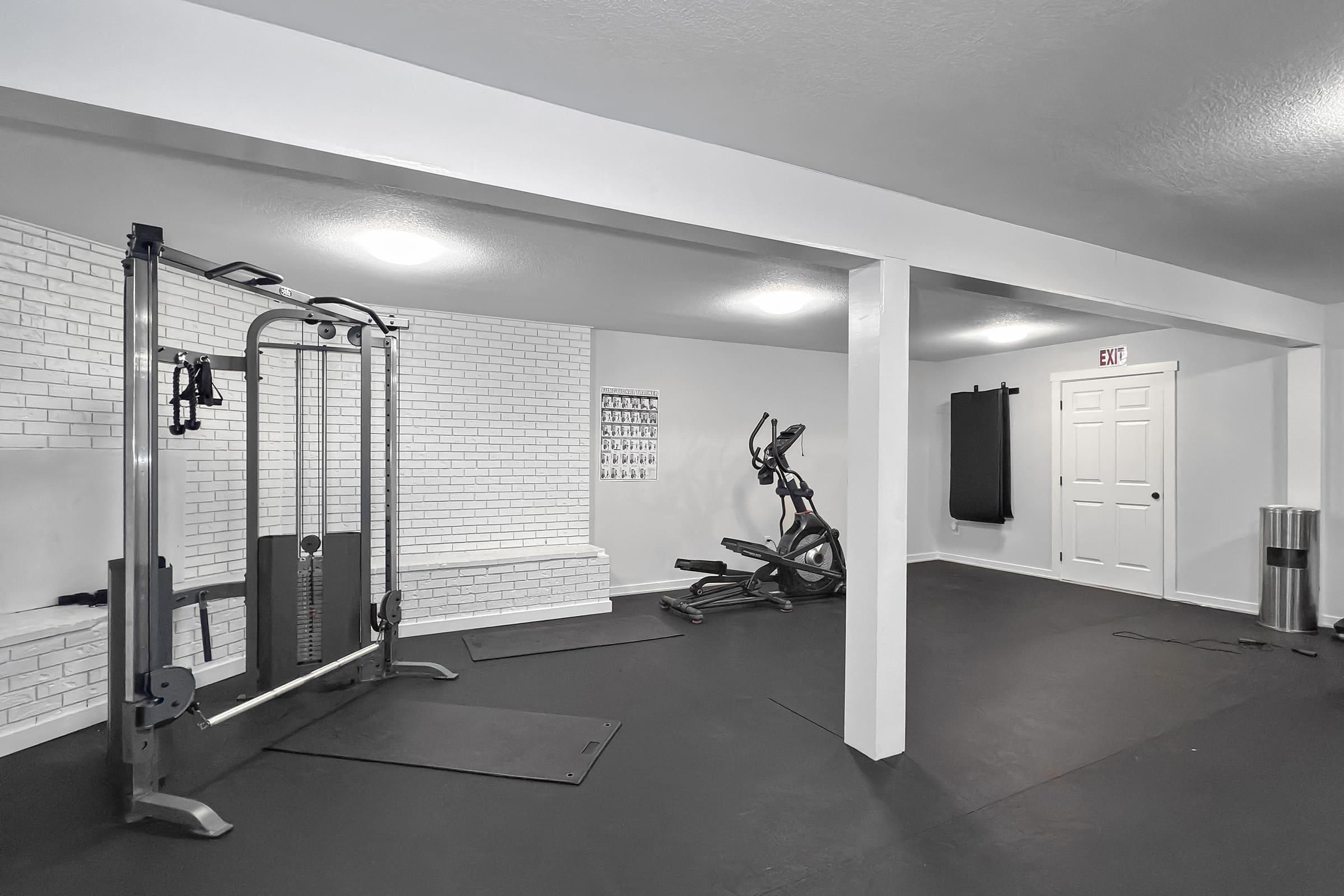 A spacious home gym featuring a cable machine, elliptical machine, and a wall with white brick design. The floor is covered with black mats, and there are large, bright lights in the ceiling. A door leads to another room, and the space is uncluttered and well-maintained.