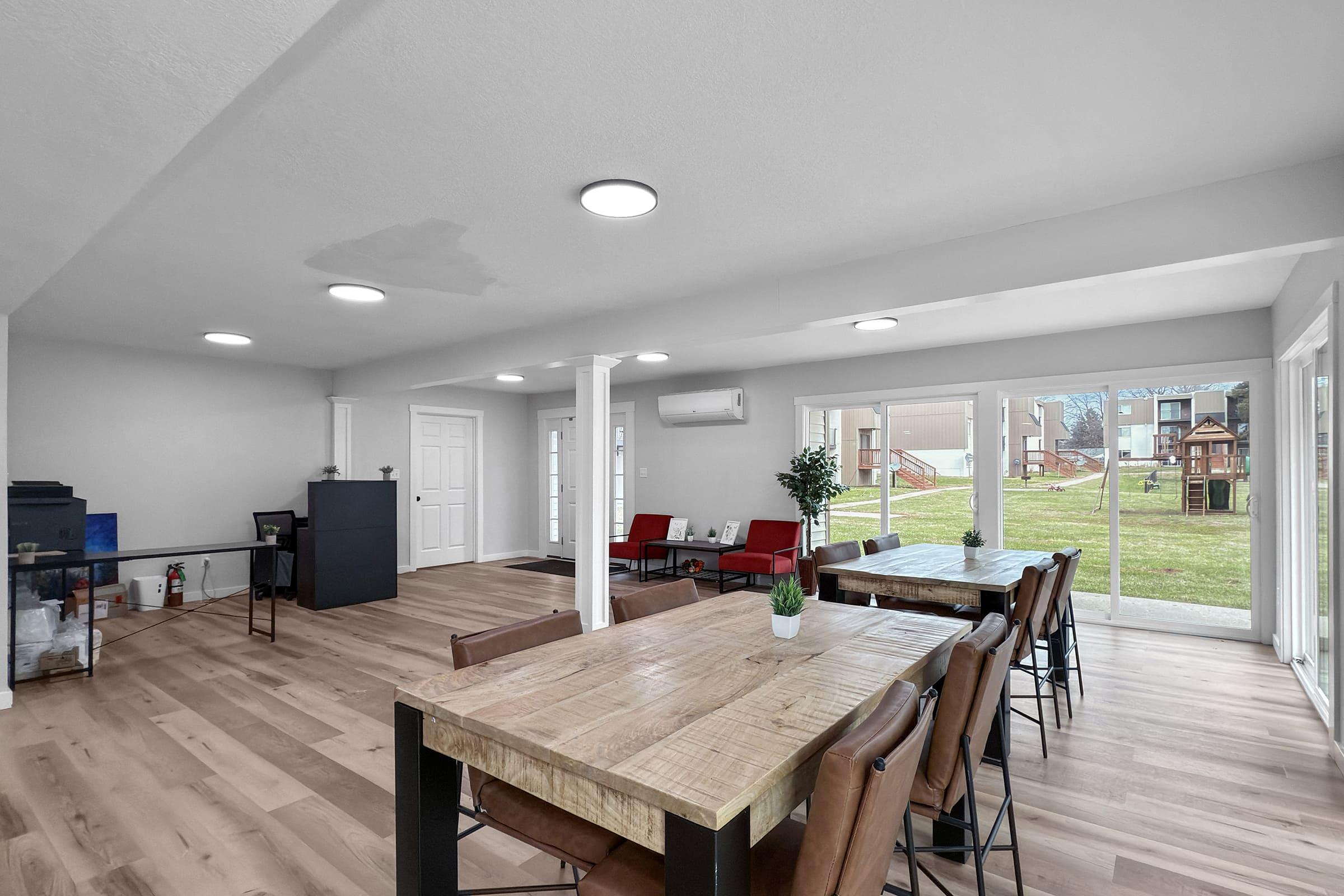 A spacious, modern interior featuring a dining area with a large wooden table and leather chairs. There are large windows allowing natural light, with views of a backyard and a playground. The room has a neutral color palette, accented by contemporary light fixtures and minimal decor.
