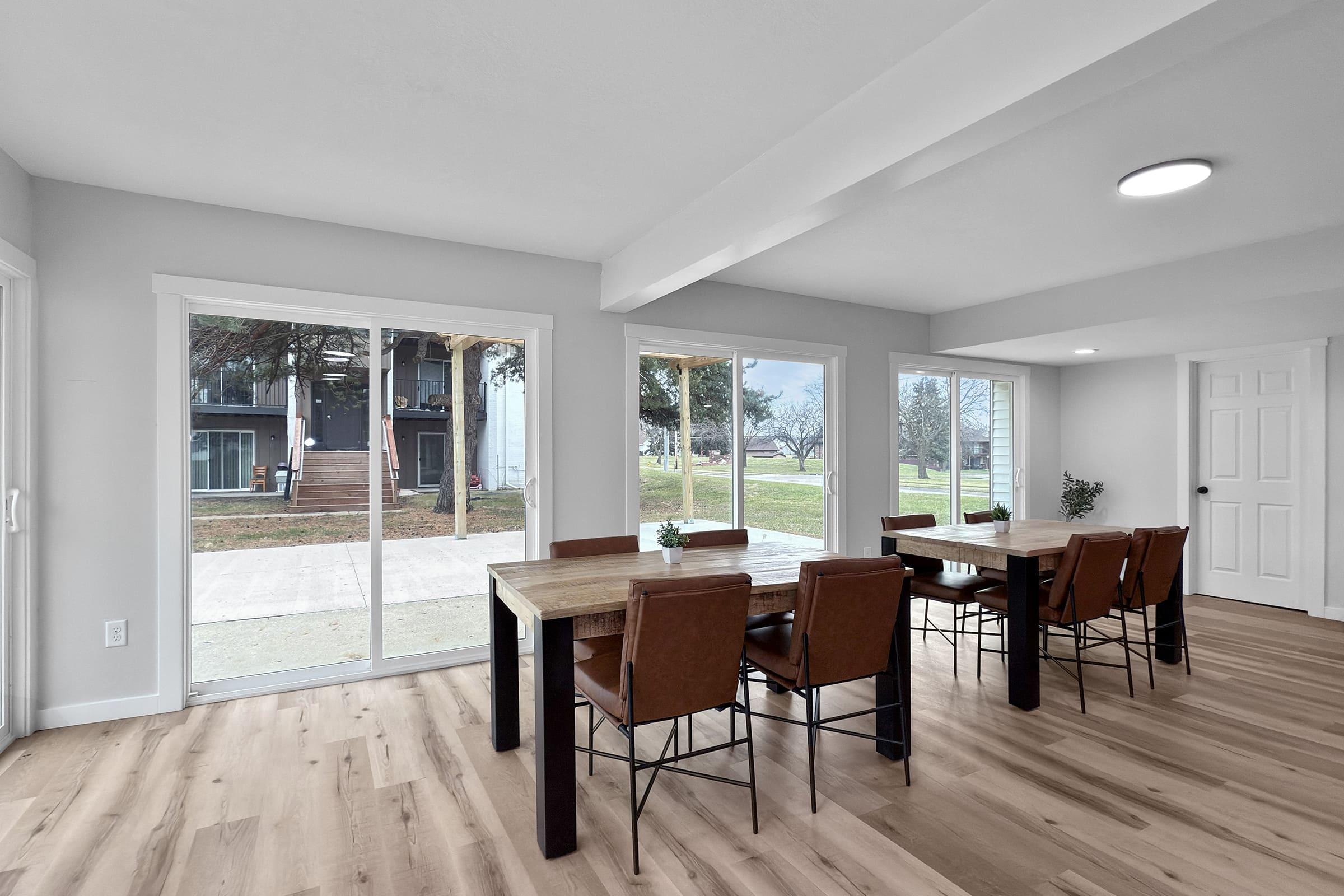 A spacious dining area featuring a large wooden table surrounded by leather chairs. Large glass sliding doors provide a view of an outdoor space with trees and a staircase. The walls are painted in light gray, and the floor is light wood, creating a modern and airy atmosphere.