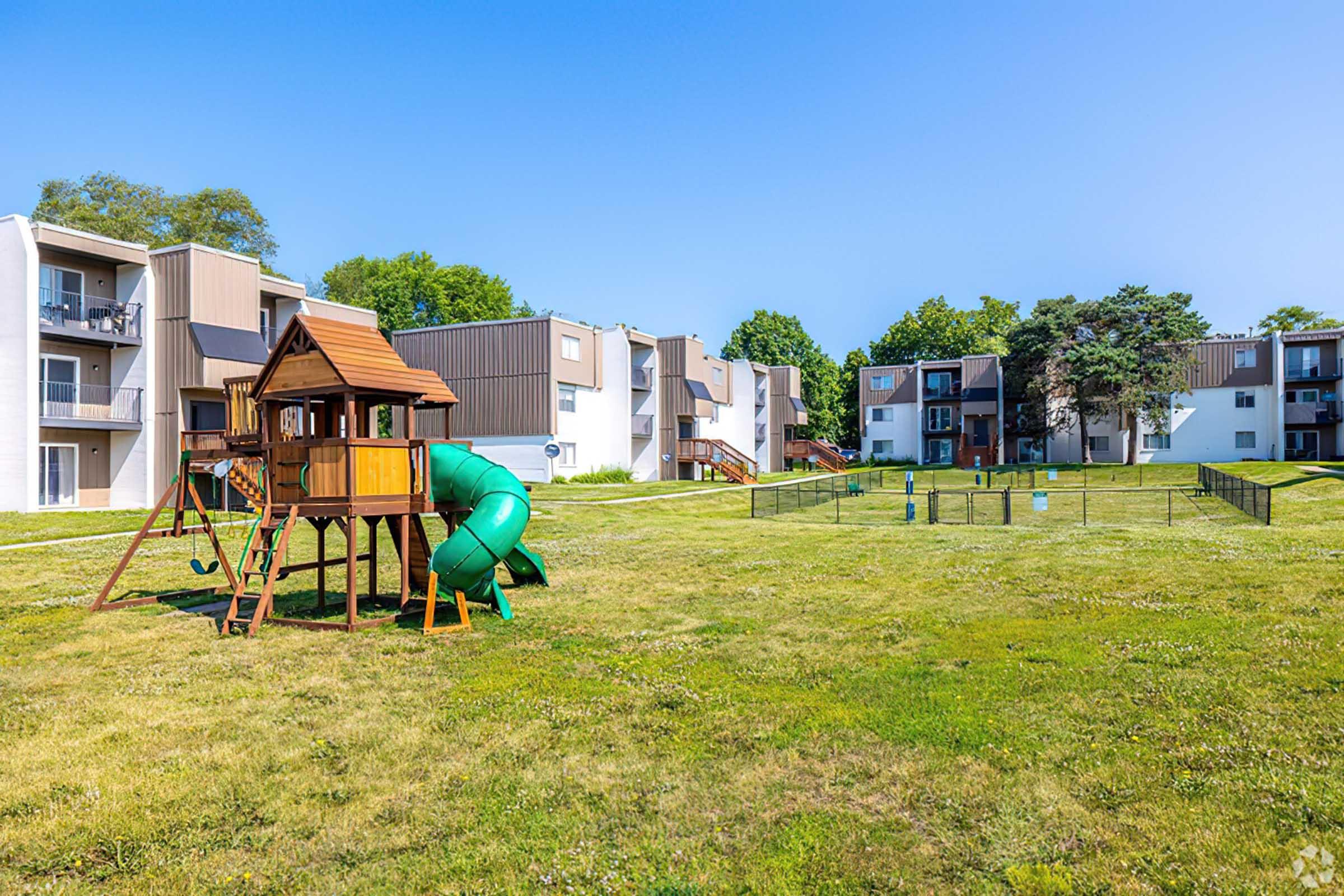 A spacious outdoor area featuring a wooden playground set with slides and swings, surrounded by grassy lawns. In the background, there are several multi-story residential buildings and some trees, creating a tranquil neighborhood atmosphere.