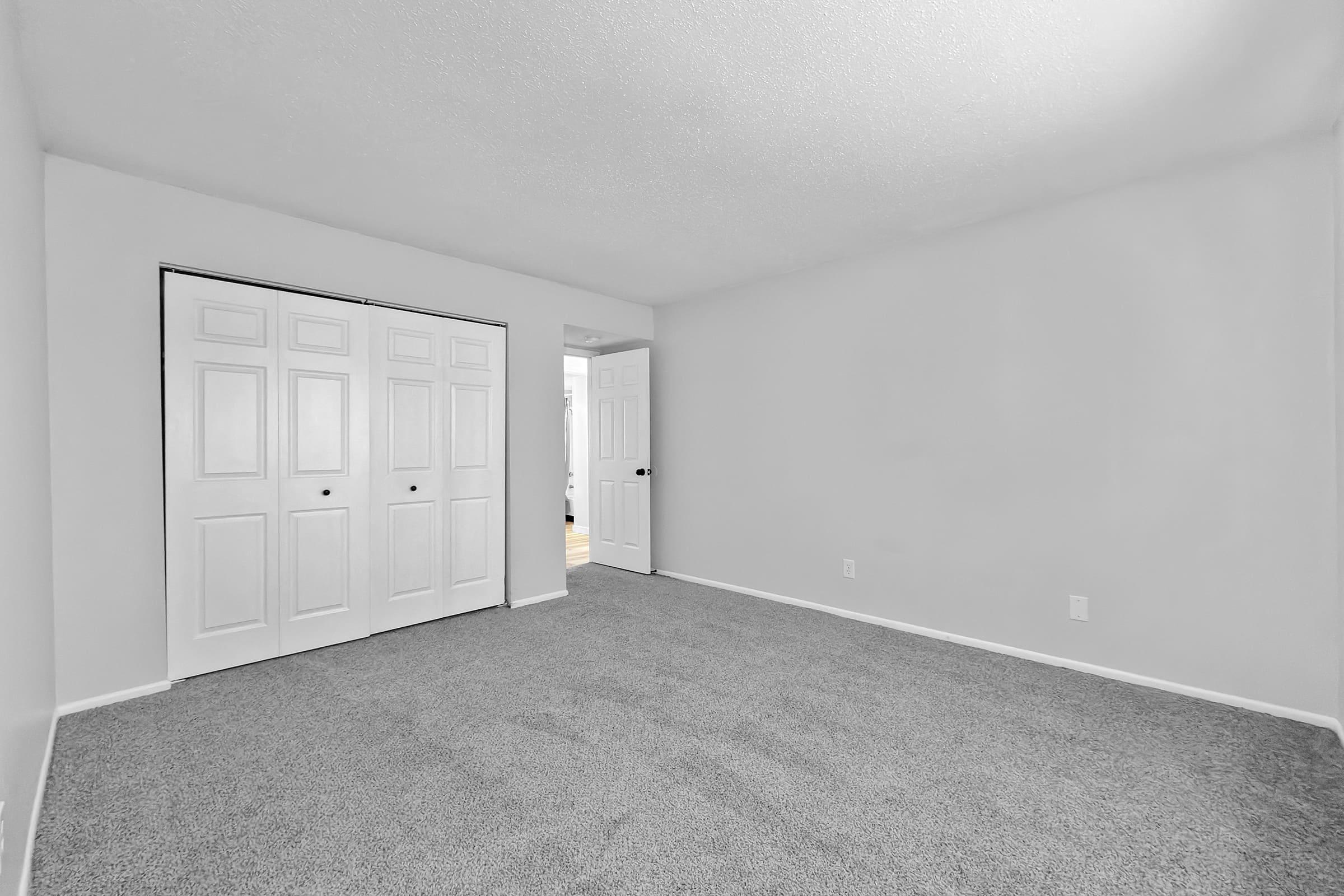 Empty room featuring light gray carpeting and plain white walls. One wall has double sliding doors, while another wall includes a doorway leading to an adjacent space. The room is brightly lit, emphasizing its spaciousness and simplicity.