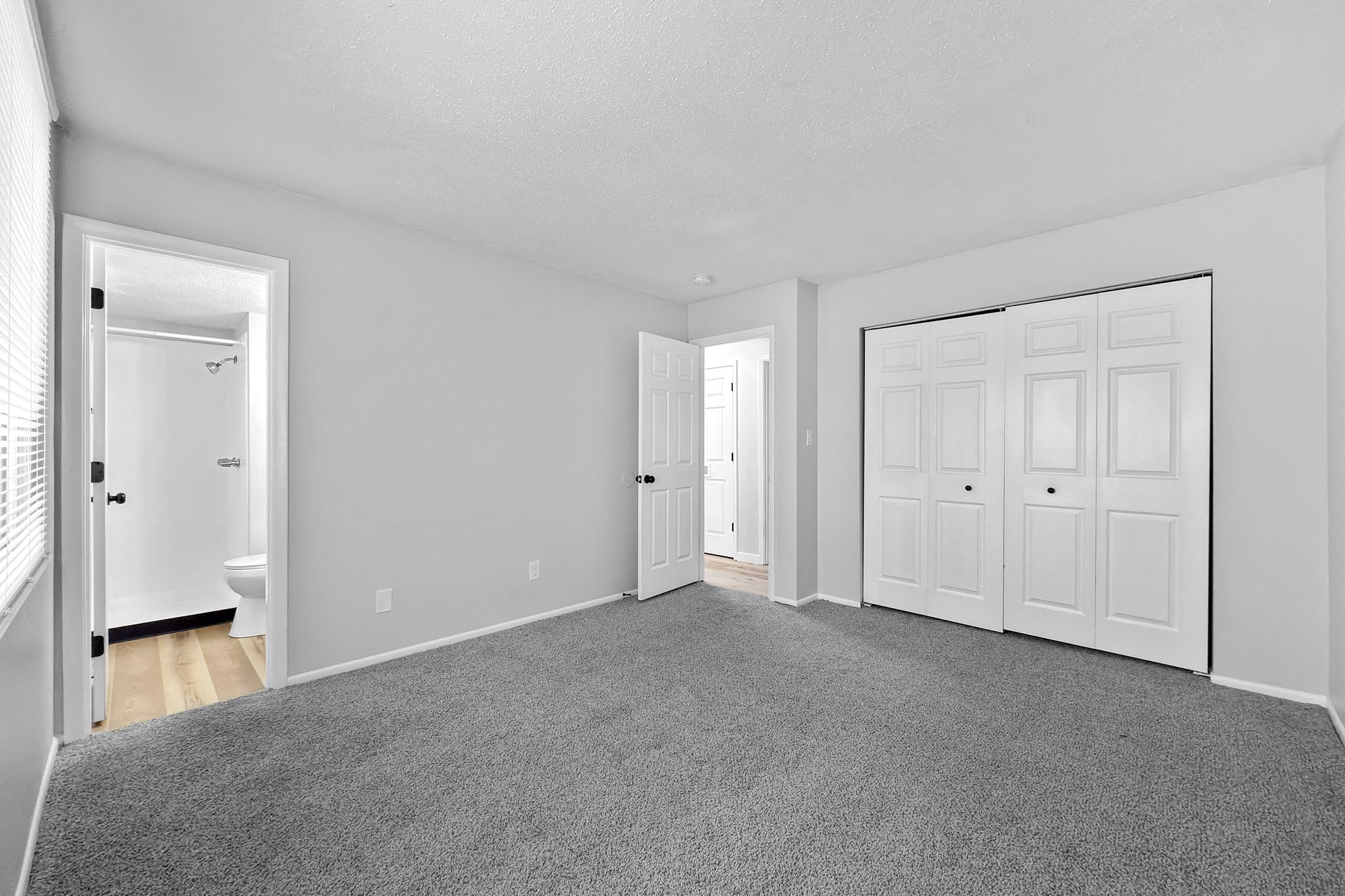 Spacious, empty room with light gray carpet, white walls, and bright natural light from a window. A bathroom door is visible on the left, while another door leads to a hallway on the right. The closet features double white doors, creating a modern and clean aesthetic.