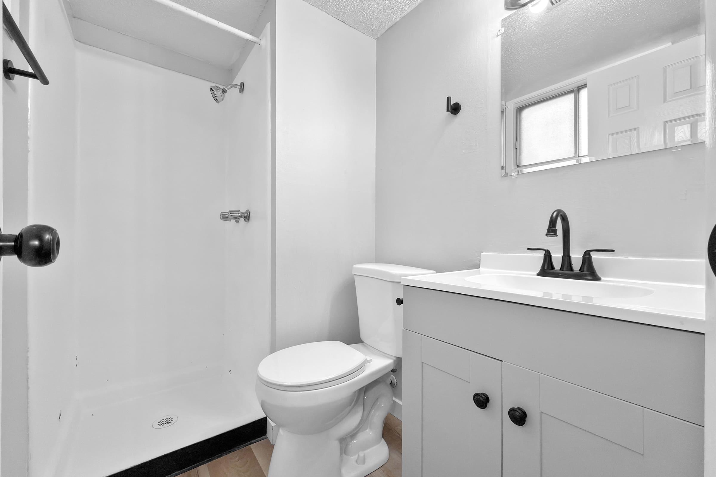 A small, modern bathroom featuring a white shower stall, a toilet, and a sink with a black faucet. The walls are painted light gray, and there is a mirror above the sink. The floor has a simple, light-colored design, and a window provides natural light.