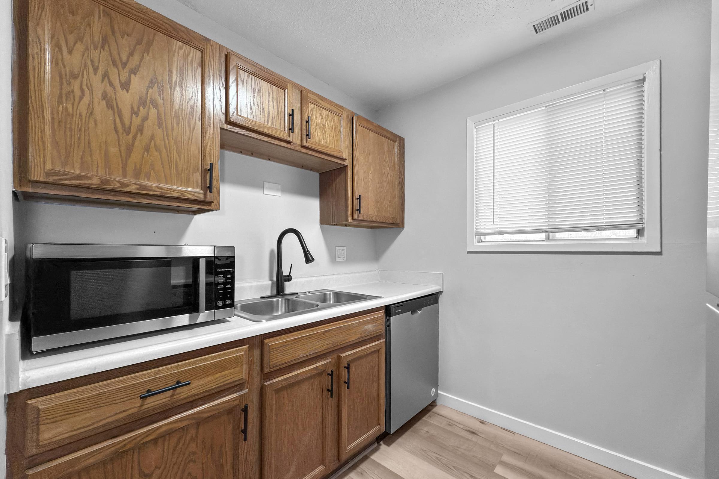 A modern kitchen featuring wooden cabinets, a stainless steel sink, a microwave, and a dishwasher. The countertop is white and there is a window with blinds providing natural light. The flooring is light-colored wood, creating a clean and inviting space.