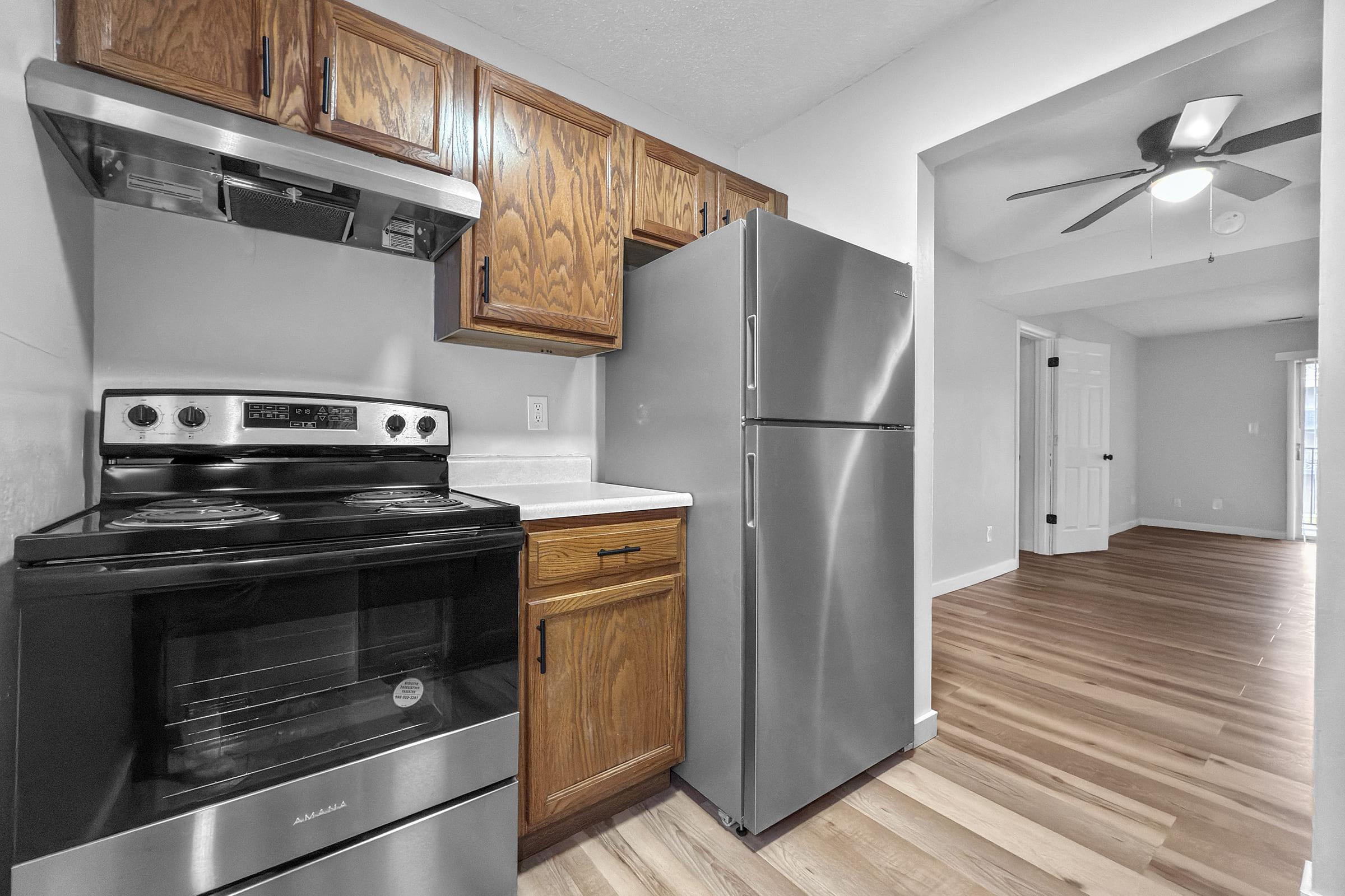 A modern kitchen featuring wooden cabinets, a stainless steel refrigerator, and a black stove. The space has light-colored walls and hardwood flooring, with an open layout leading to a bright living area. A ceiling fan is visible in the room beyond.