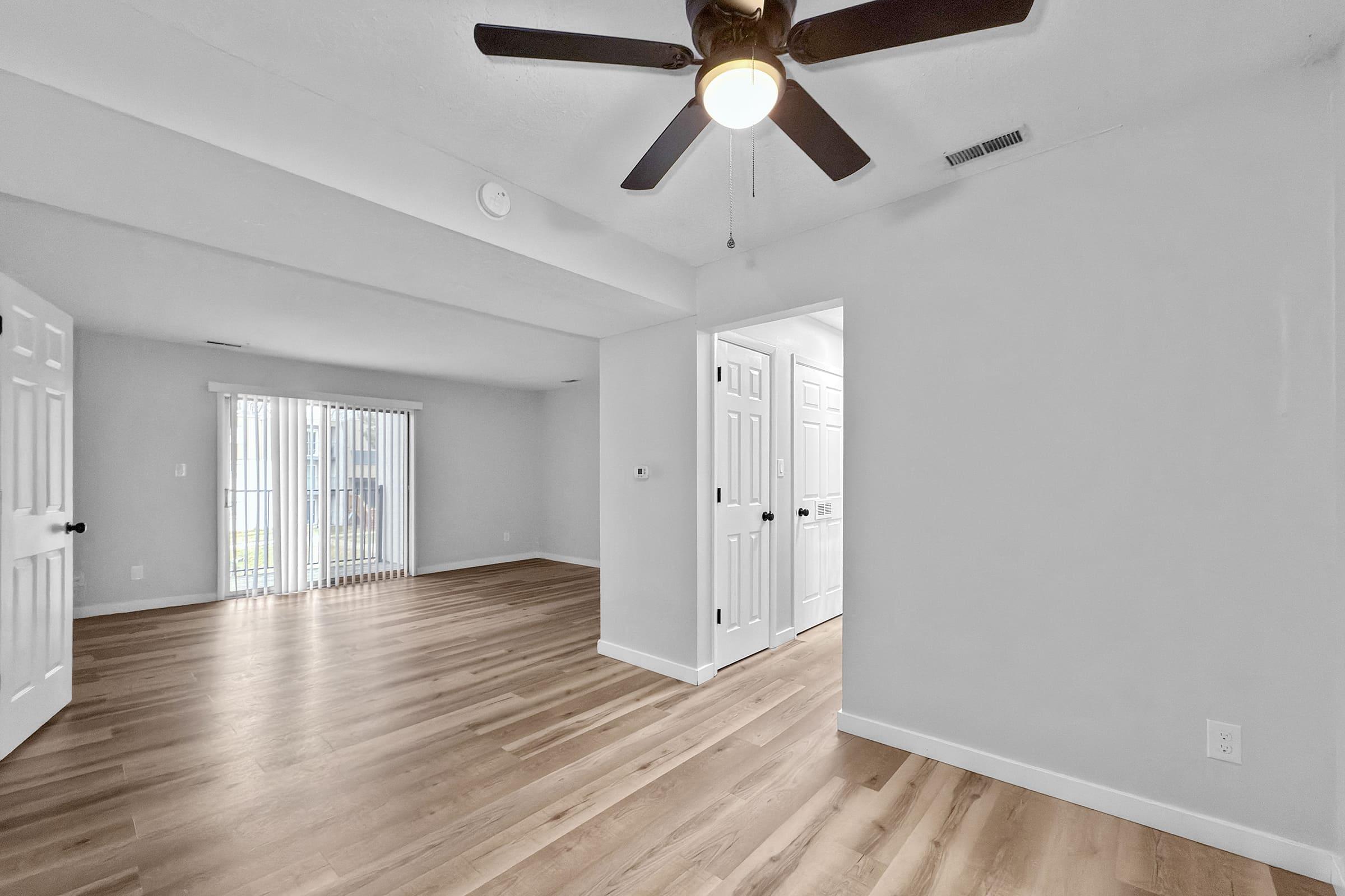 Spacious interior view of a newly renovated room featuring light-colored walls, hardwood flooring, and a ceiling fan. A doorway leads to a closet on one side, while large sliding glass doors provide access to natural light from outside. The overall design is modern and minimalist.