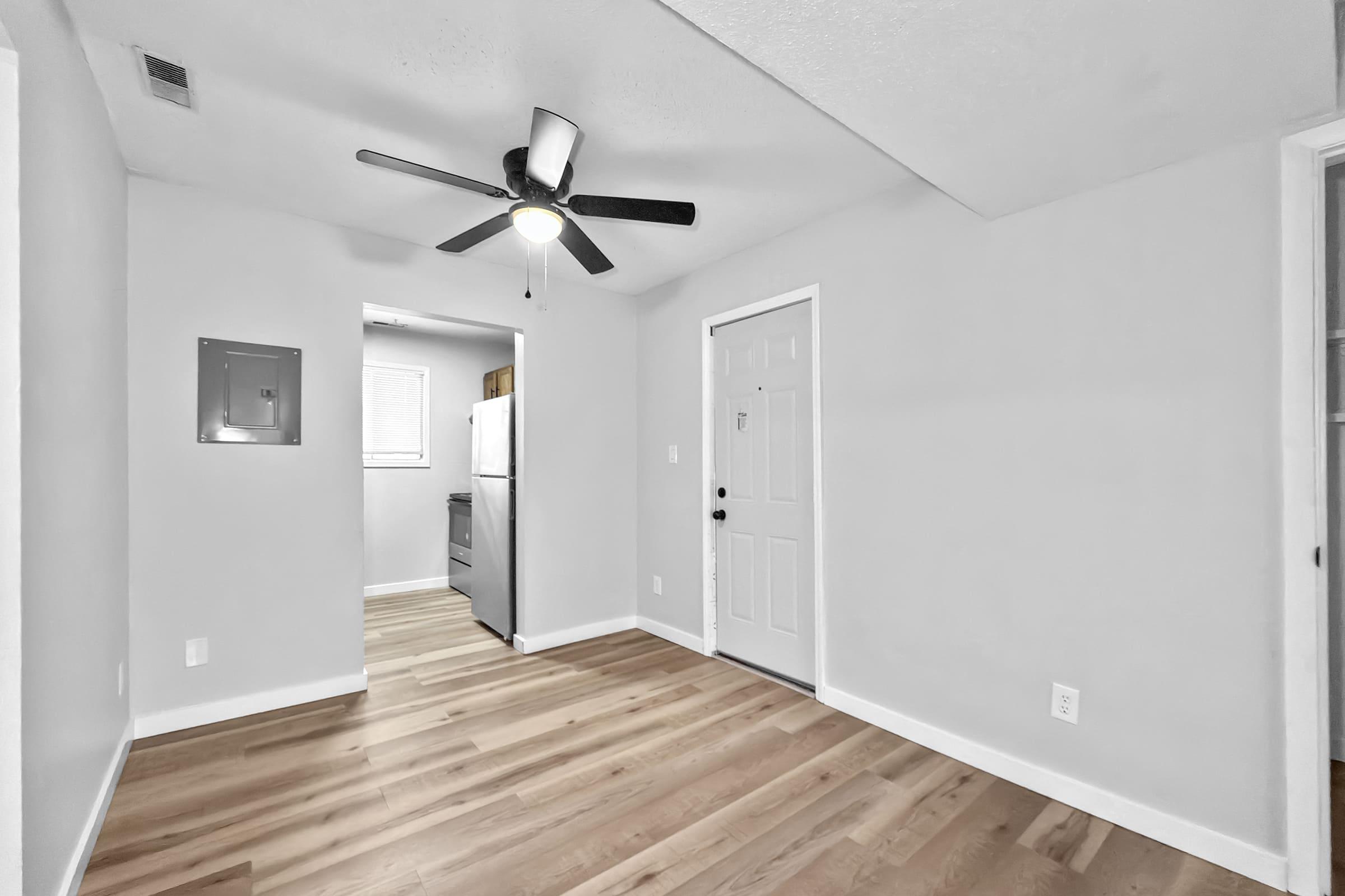 Interior view of a room featuring light gray walls, a ceiling fan, and laminate flooring. A doorway leads to a kitchen area with a refrigerator visible. A utility panel is mounted on the wall, and there’s a door leading outside. Natural light comes from a window in the adjacent room.