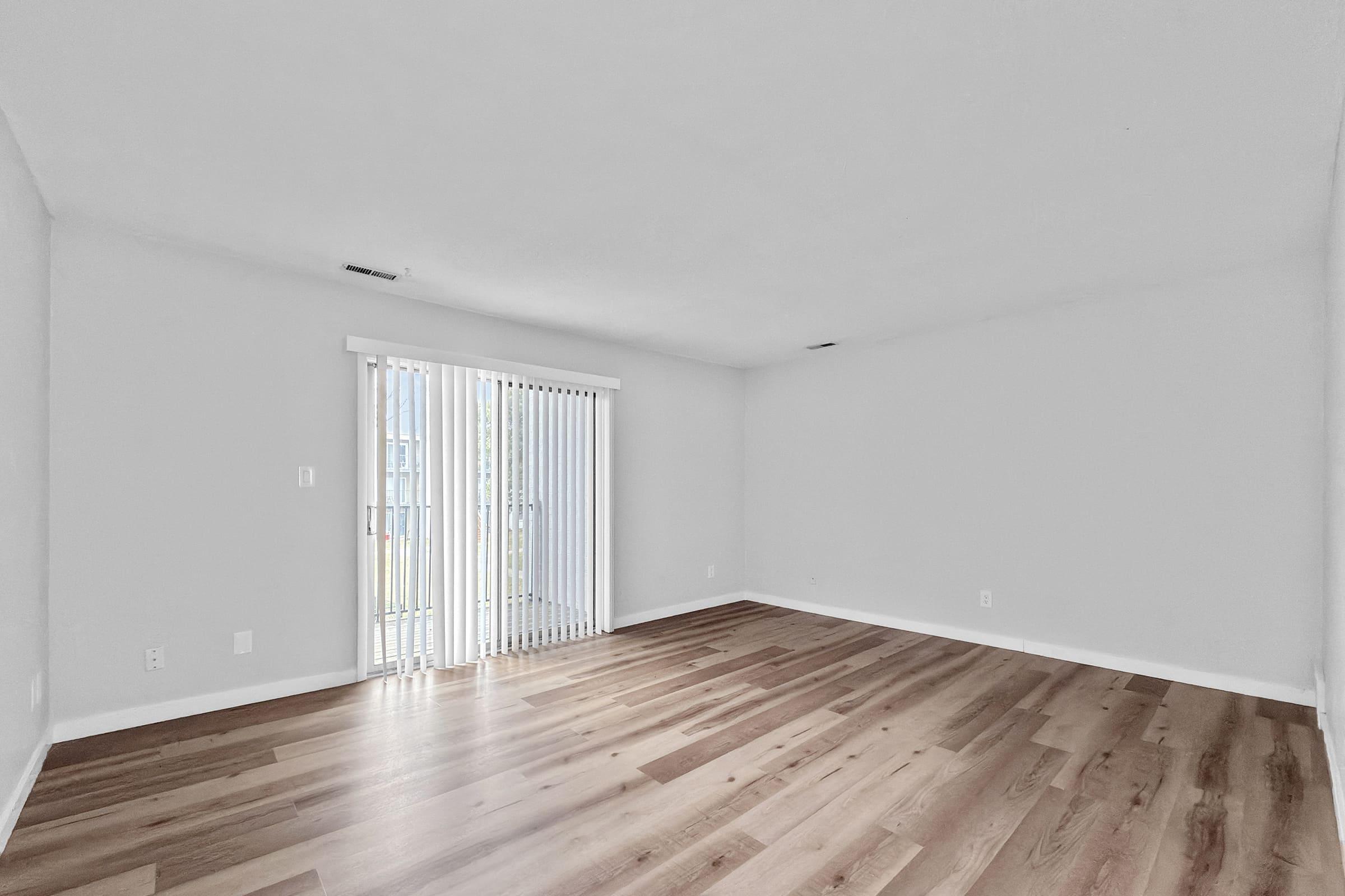 Empty room with light gray walls and wooden flooring. Large sliding glass door with vertical blinds leads to an outdoor area. The space is well-lit with natural light, creating a bright and airy atmosphere.