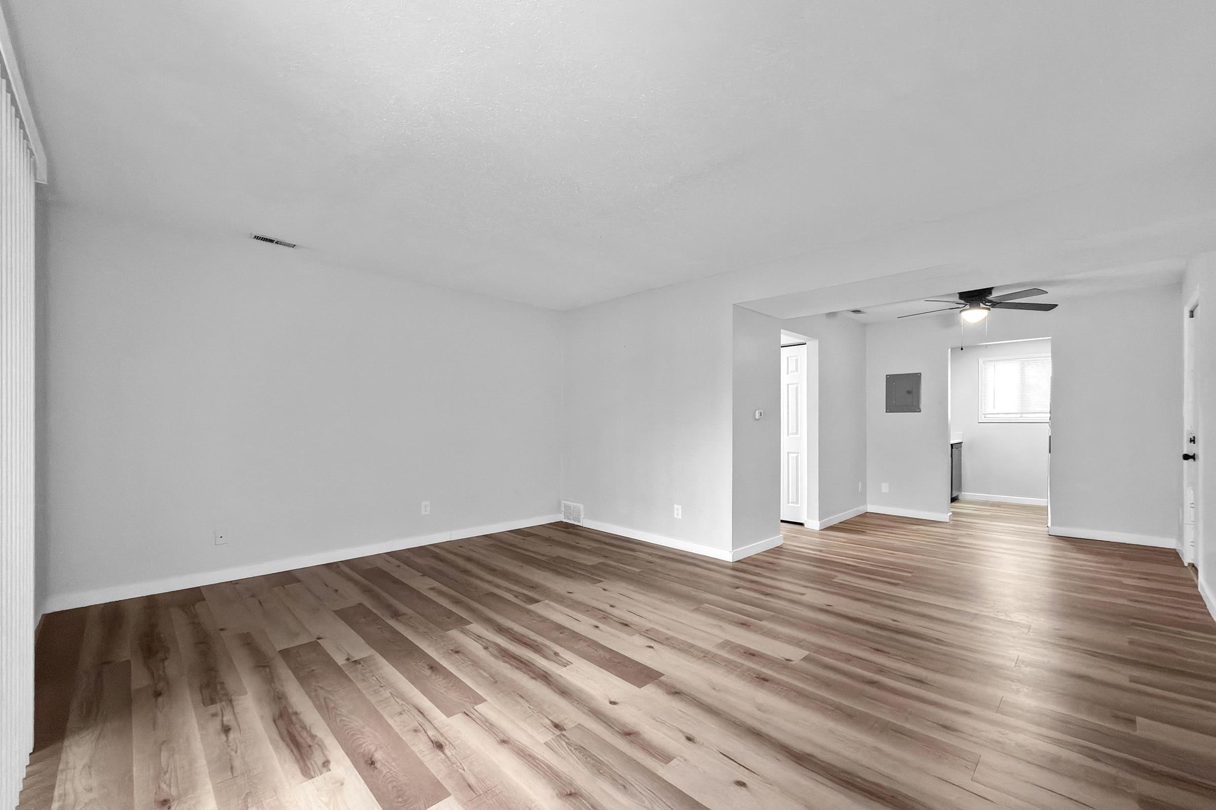 A spacious, empty living room with light hardwood flooring and white walls. There is a doorway leading to another room, and a ceiling fan visible. Natural light enters through a window, creating a bright and airy atmosphere.
