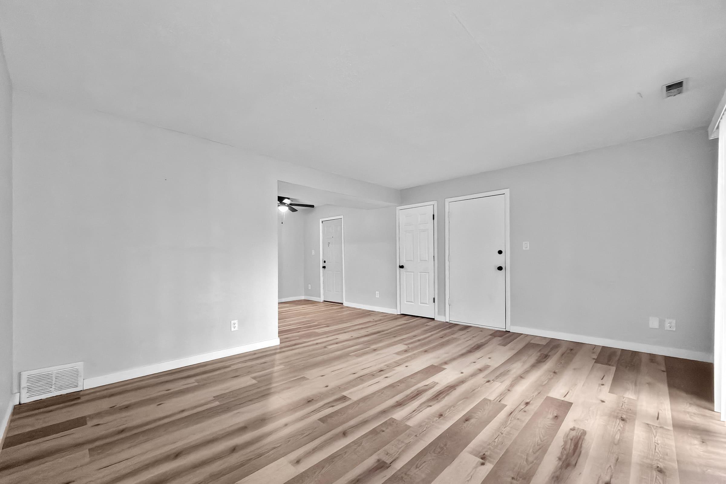 A spacious, empty room with light-colored walls and smooth, wooden flooring. There are two closed doors along one wall and a ceiling fan visible in the corner. The room has ample natural light, creating a bright and airy atmosphere, suitable for various interior design possibilities.