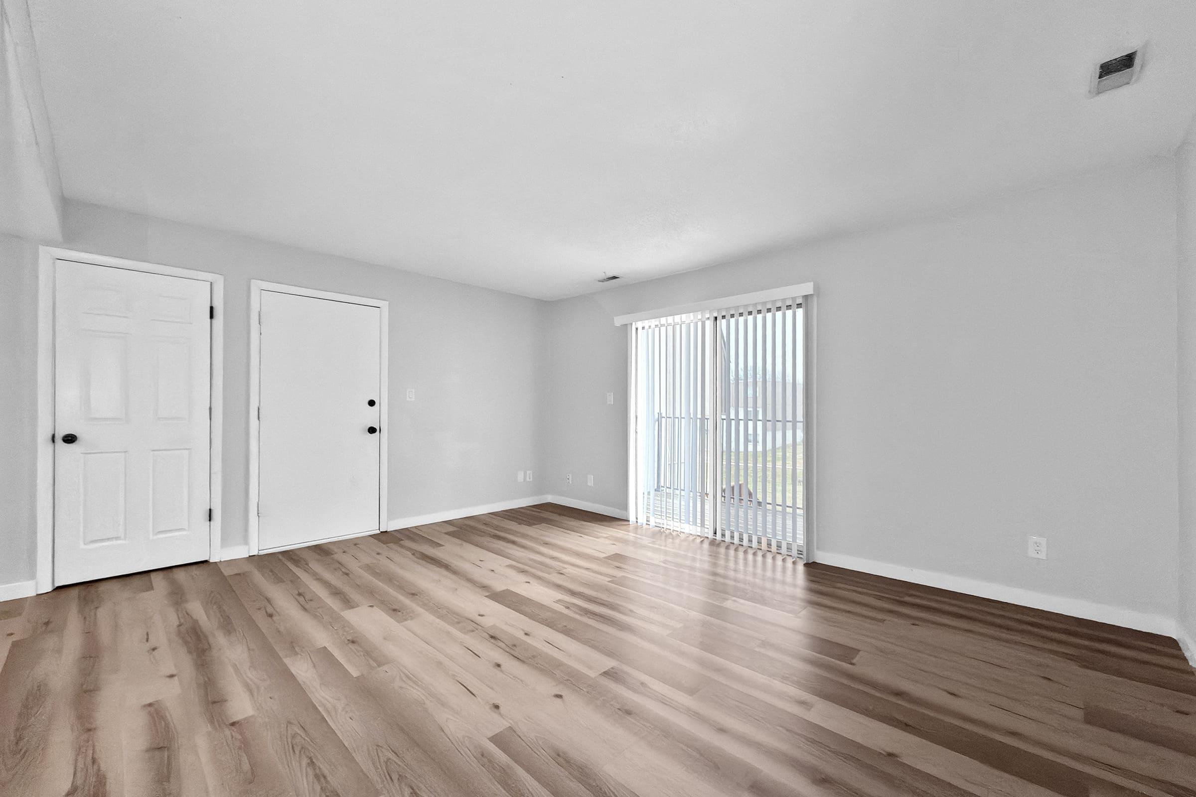 A spacious, empty room with light grey walls and a wooden floor. There are two doors leading to closets or rooms, and a large window with vertical blinds allowing natural light to enter. The room appears bright and inviting, suitable for various decorating options.